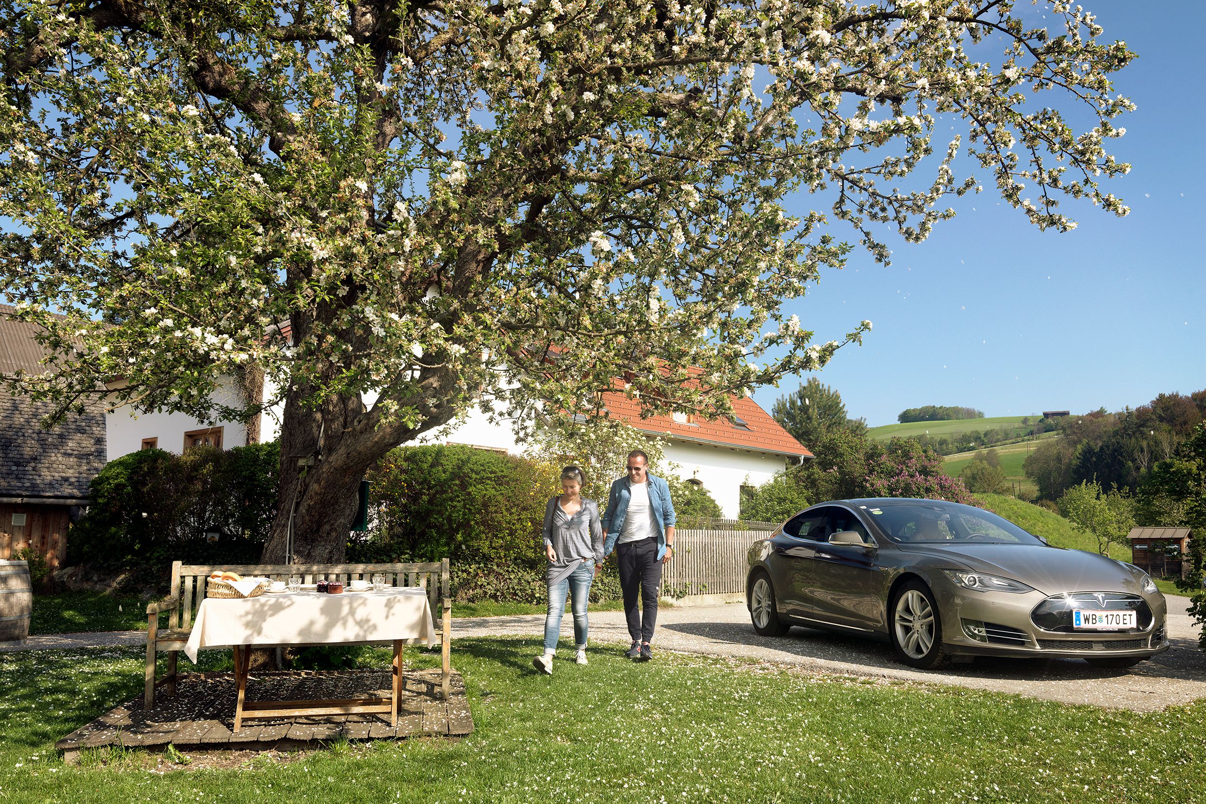 Pár se prochází vedle zaparkovaného elektromobilu pod rozkvetlým stromem ve venkovské zahradě.