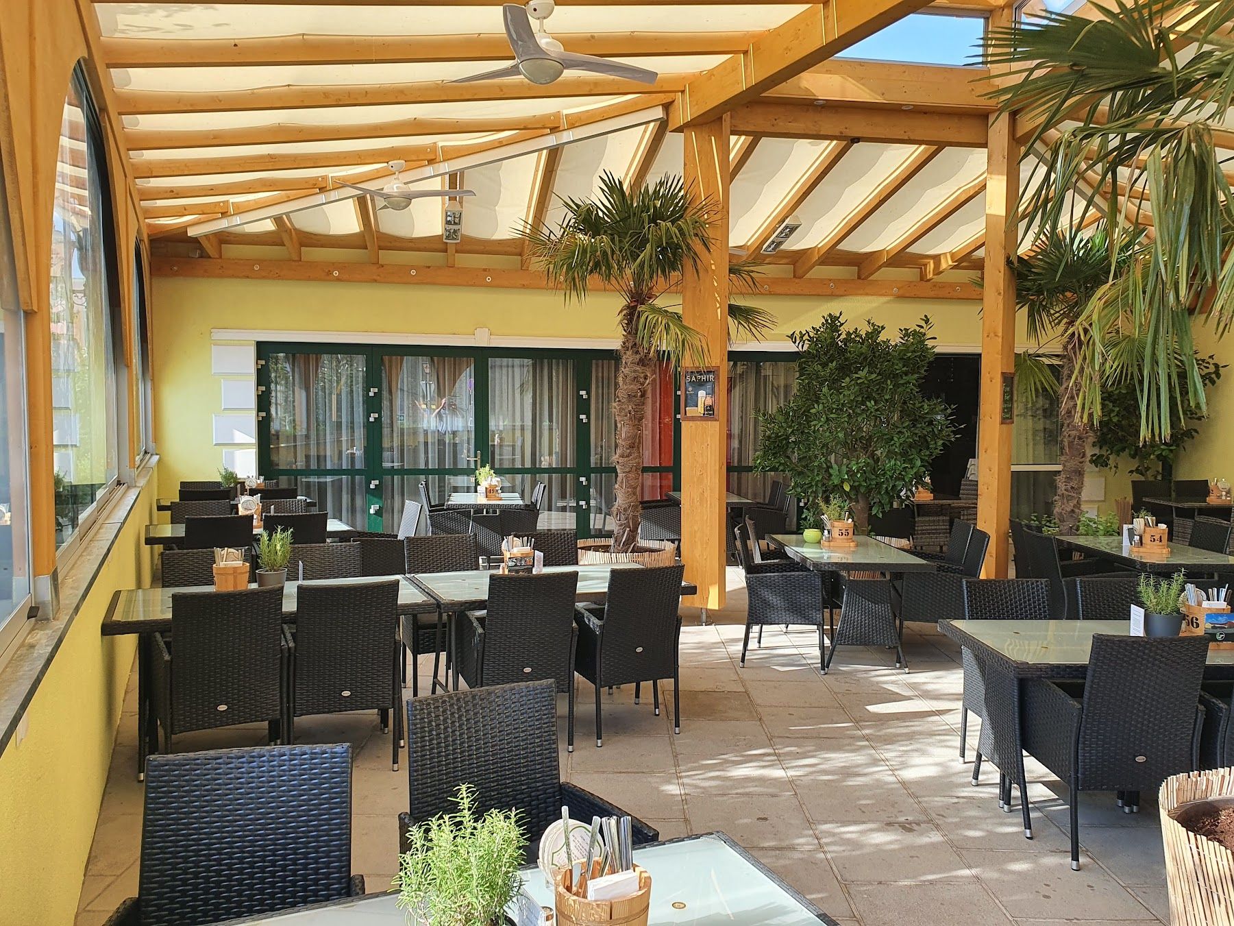 Krytá terasa restaurace s černým ratanovým nábytkem a rostlinami.