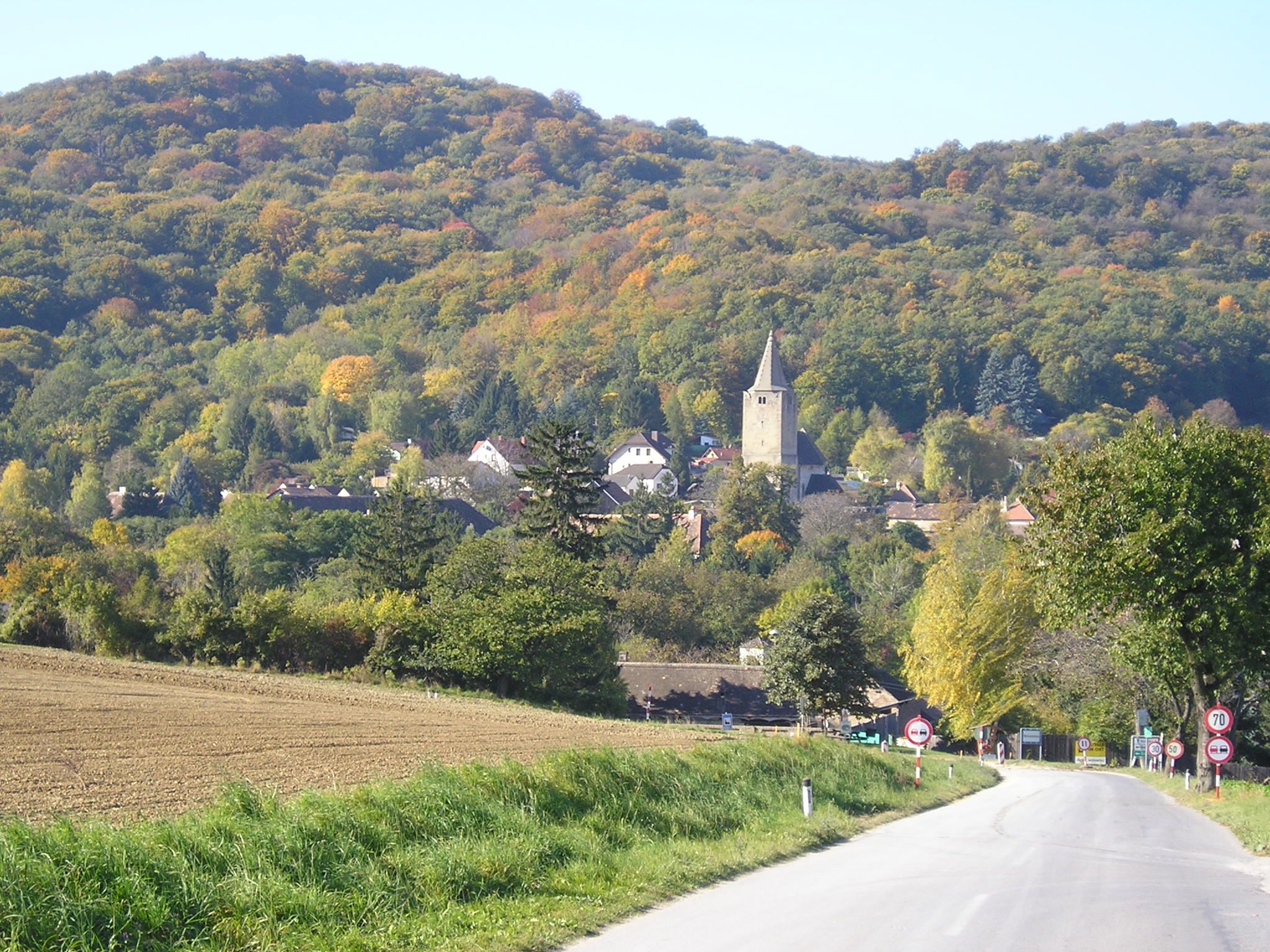 Malebná krajina se silnicí vedoucí do vesnice s kostelem, obklopená zalesněnými kopci na podzim.