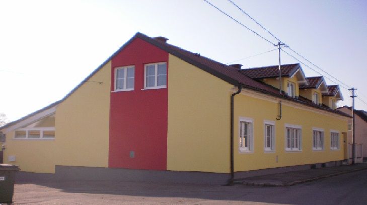 Vnější pohled na žlutočervenou budovu s několika okny a šikmou střechou.
