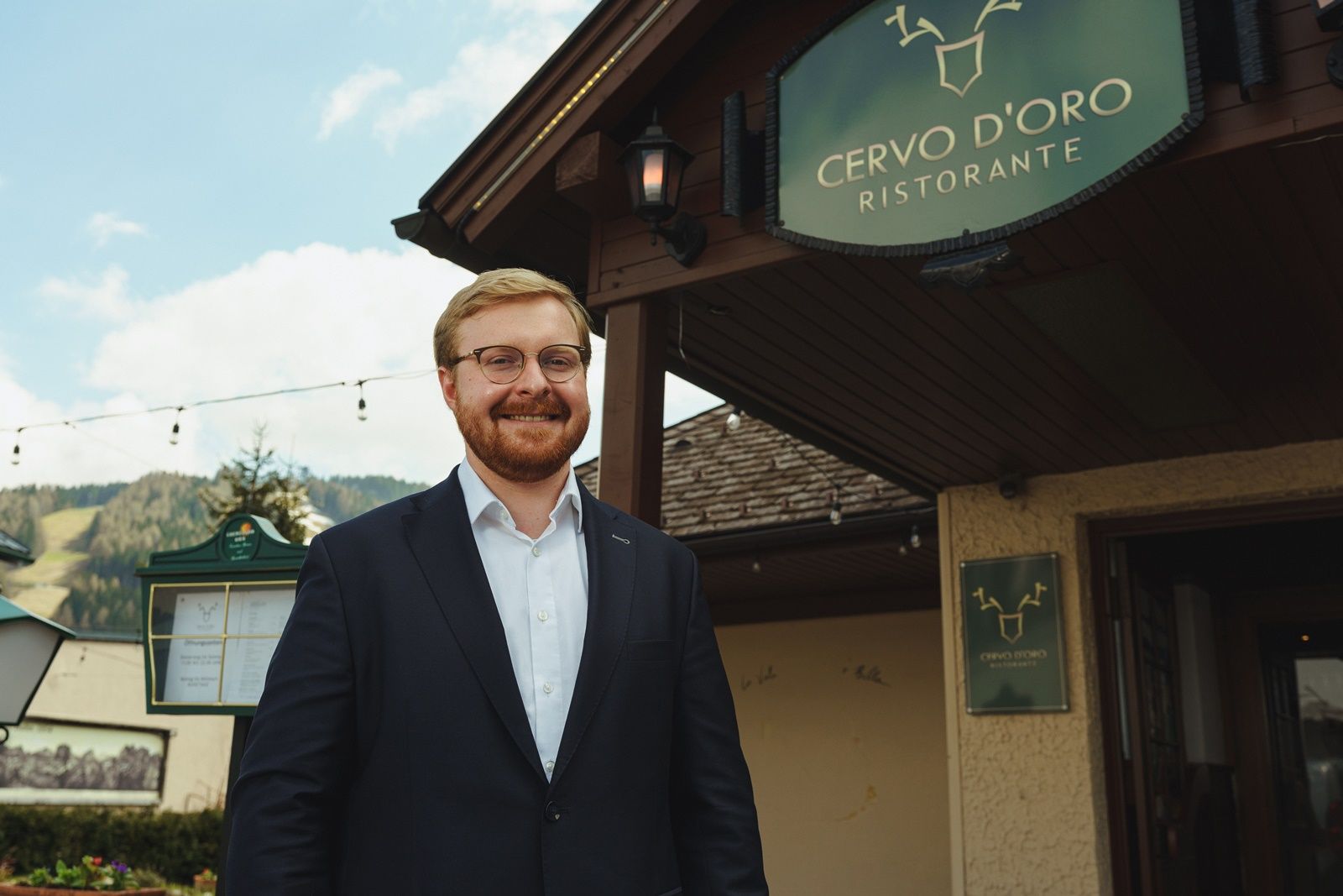 Před restaurací Cervo D'Oro stojí usměvavý muž v obleku.