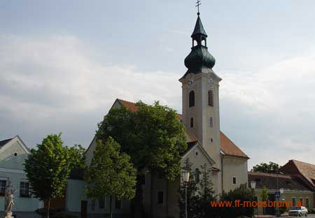 Kostel v Moosbrunnu se zelenou kostelní věží a okolními budovami.