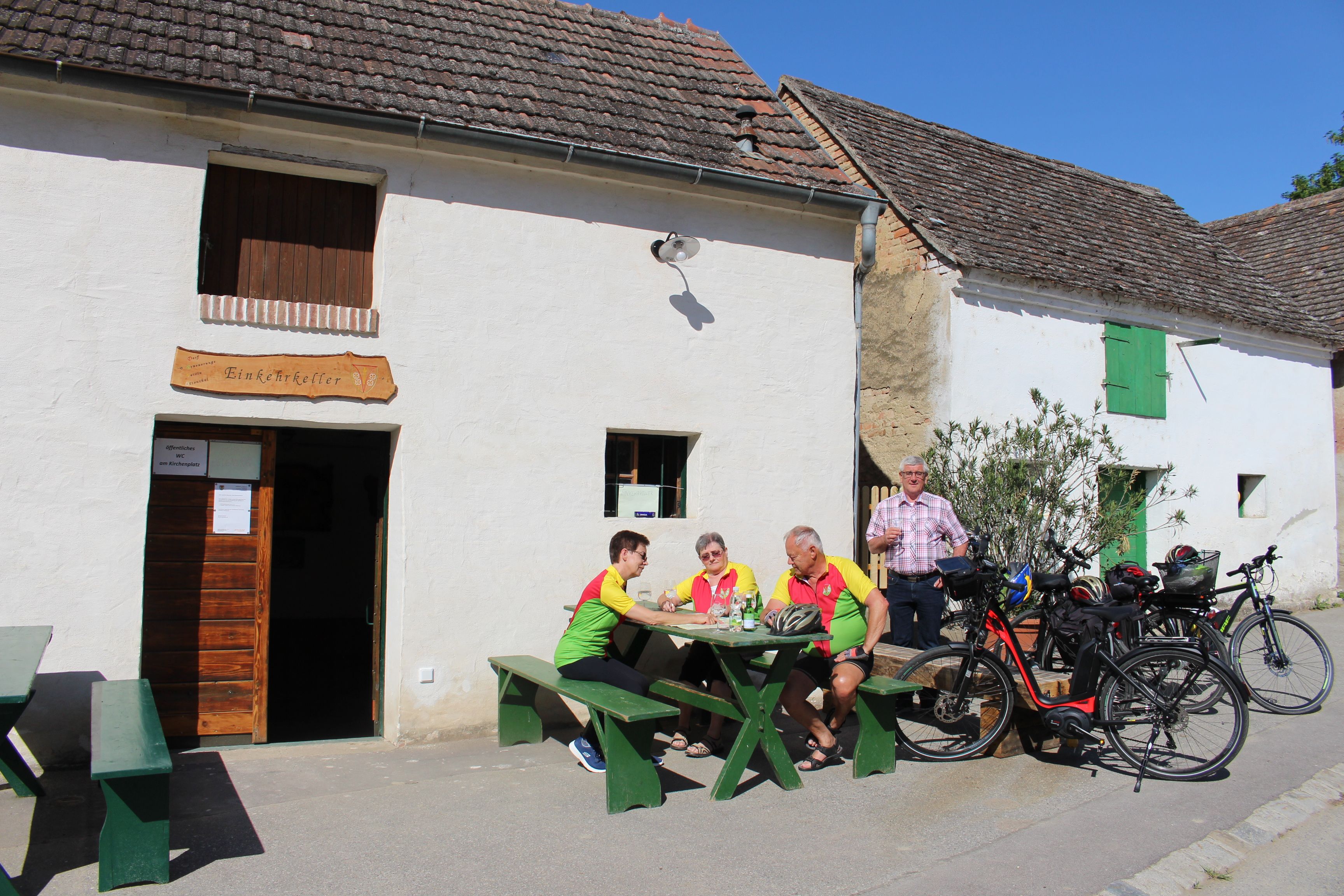 Skupina lidí sedí u stolu před restaurací „Einkehrkeller Ottenthal“. Vedle nich jsou zaparkovaná kola. Je slunečný den.