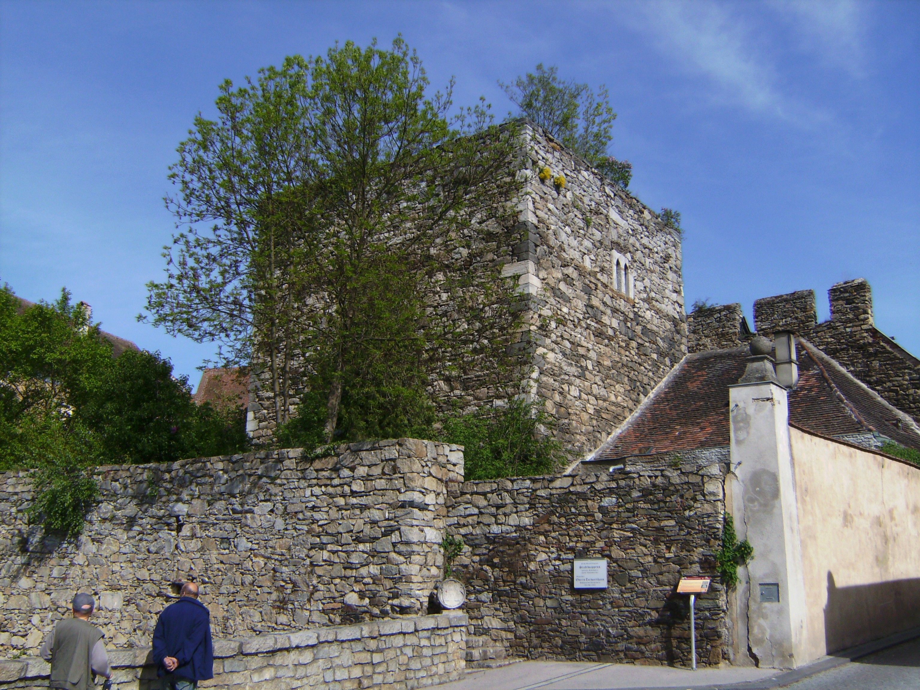 Historická kamenná zeď a věž v Drosendorf-Zissersdorfu, obklopená stromy a modrou oblohou.