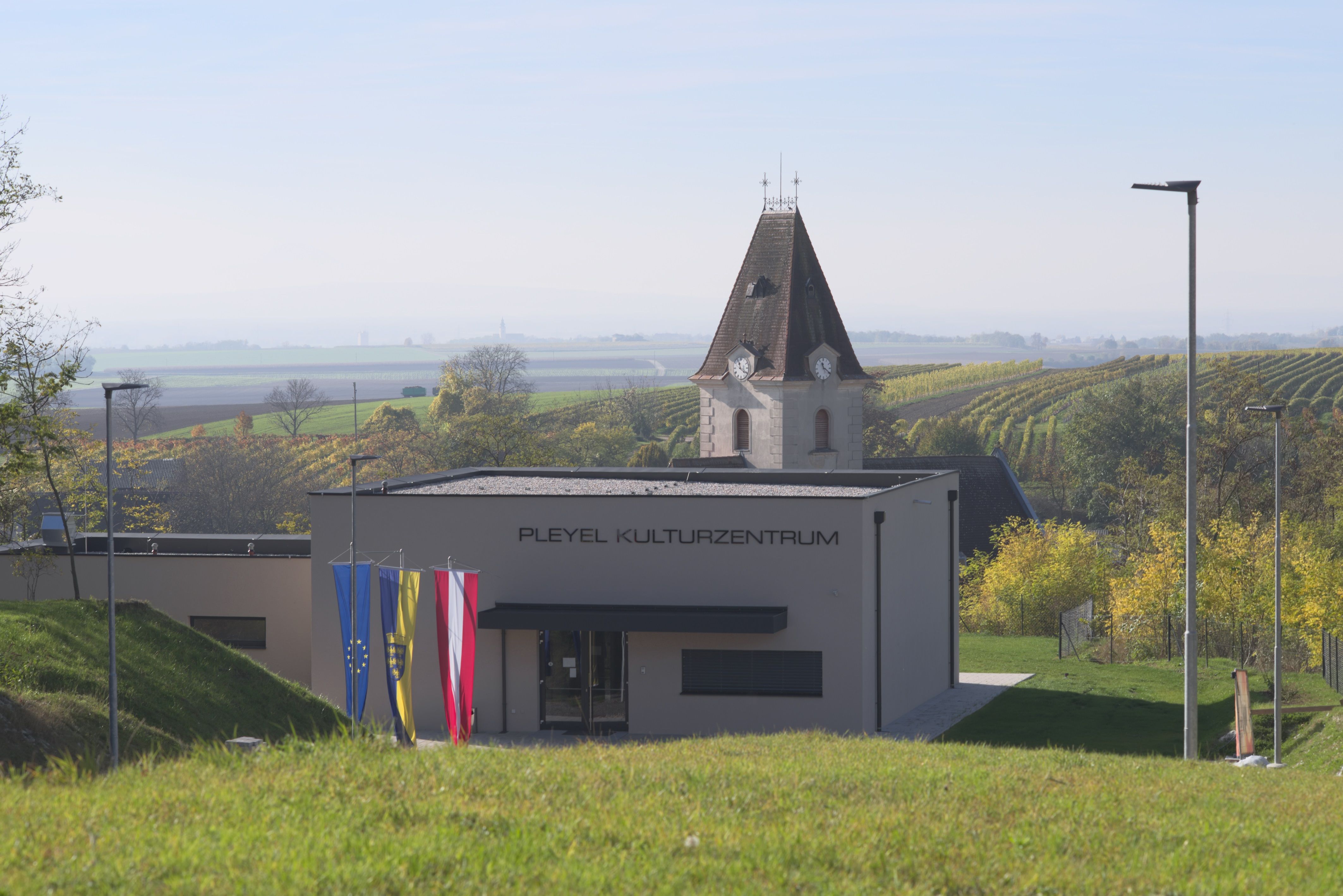 Moderní budova s nápisem "Pleyel Cultural Centre" před venkovskou krajinou s vinicemi a kostelní věží.
