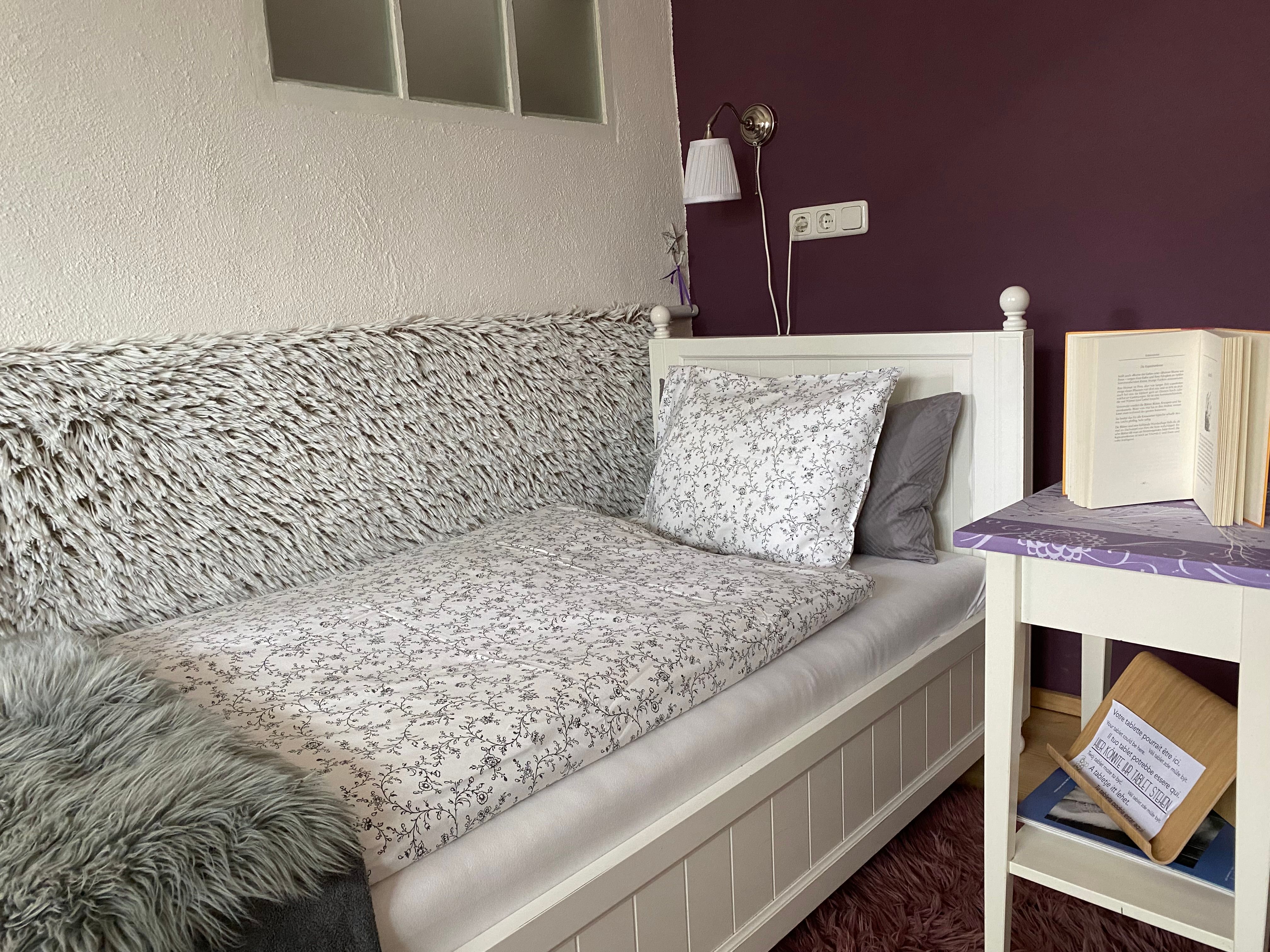 Útulná ložnice s jednolůžkem, fialovou stěnou, nočním stolkem s knihou a lampou.