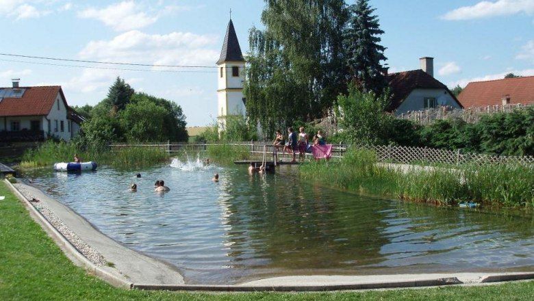 Přírodní koupací jezírko s koupajícími se lidmi. V pozadí je vidět kostelní věž a domy.