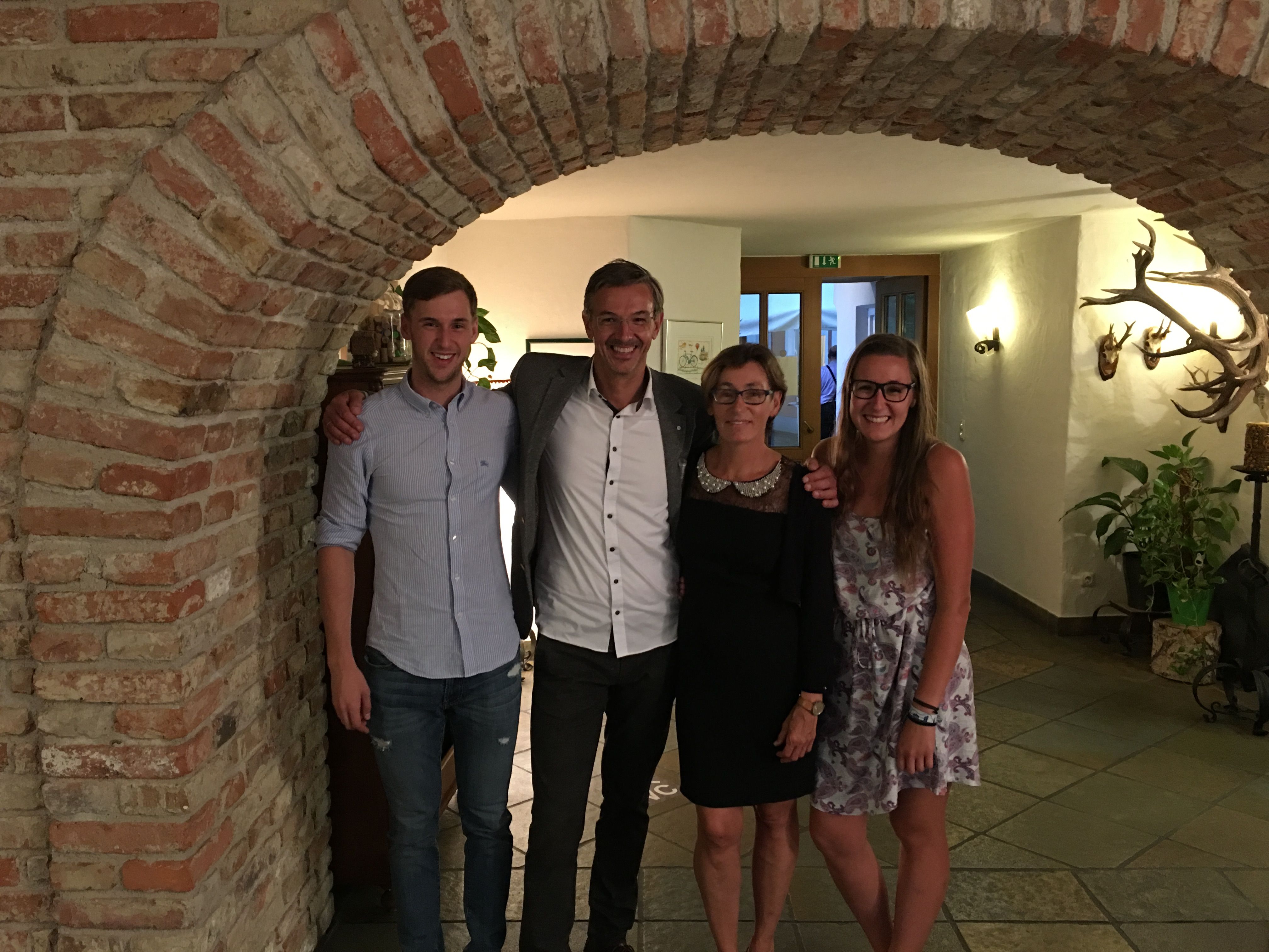 Čtyři lidé stojí s úsměvem pod cihlovým obloukem v místnosti s loveckými trofejemi na stěně.