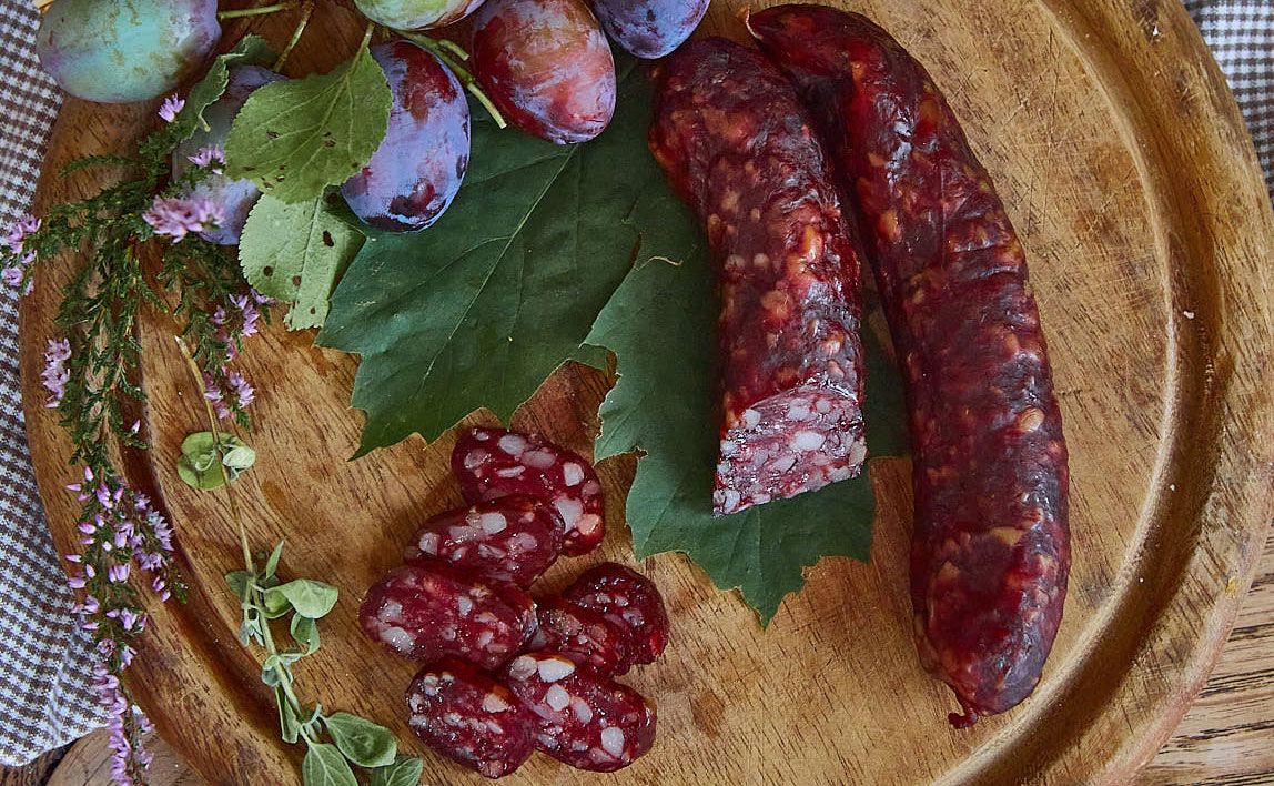 Kaminwurzn a švestky na dřevěné desce zdobené zelenými listy a bylinkami.