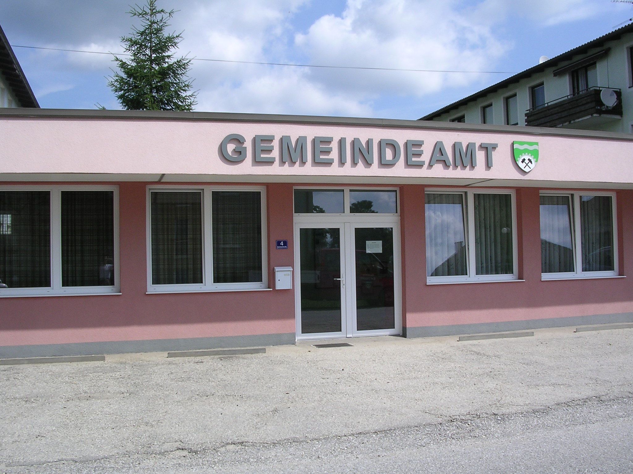 Pohled zvenčí na růžovou budovu s nápisem "GEMEINDEAMT" a erbem.