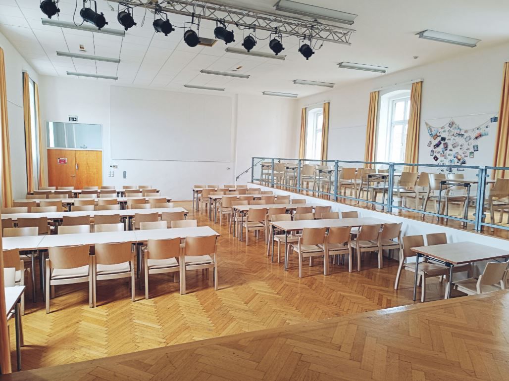Prázdný sál s dřevěnou podlahou, stoly a židlemi, pódiem a osvětlením na stropě.
