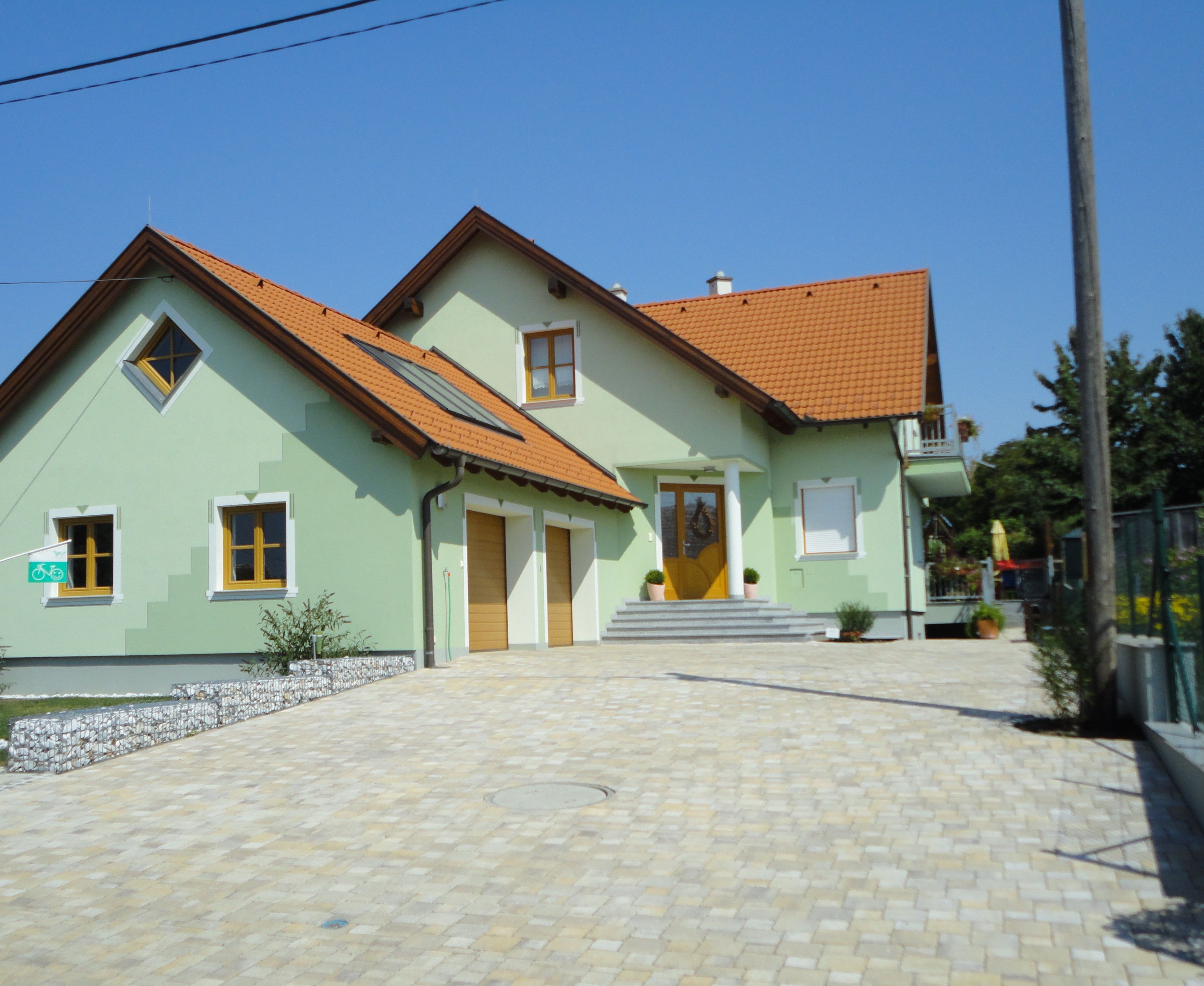 Moderní dvoupatrový dům se zeleným nátěrem a červenými střešními taškami, obklopený dlážděným dvorem.