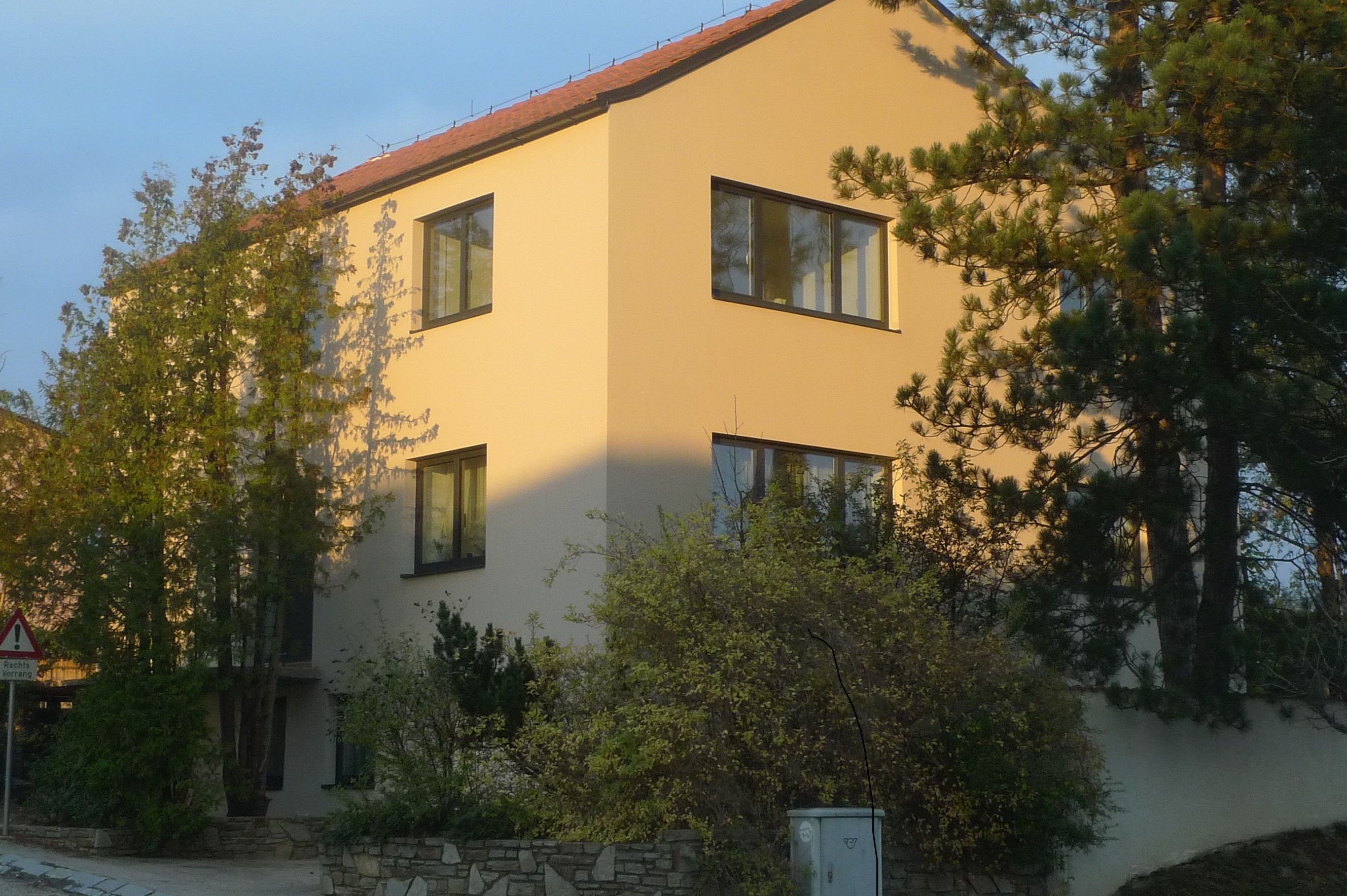 Moderní třípodlažní dům se žlutou fasádou a červenými střešními taškami, obklopený stromy a keři.