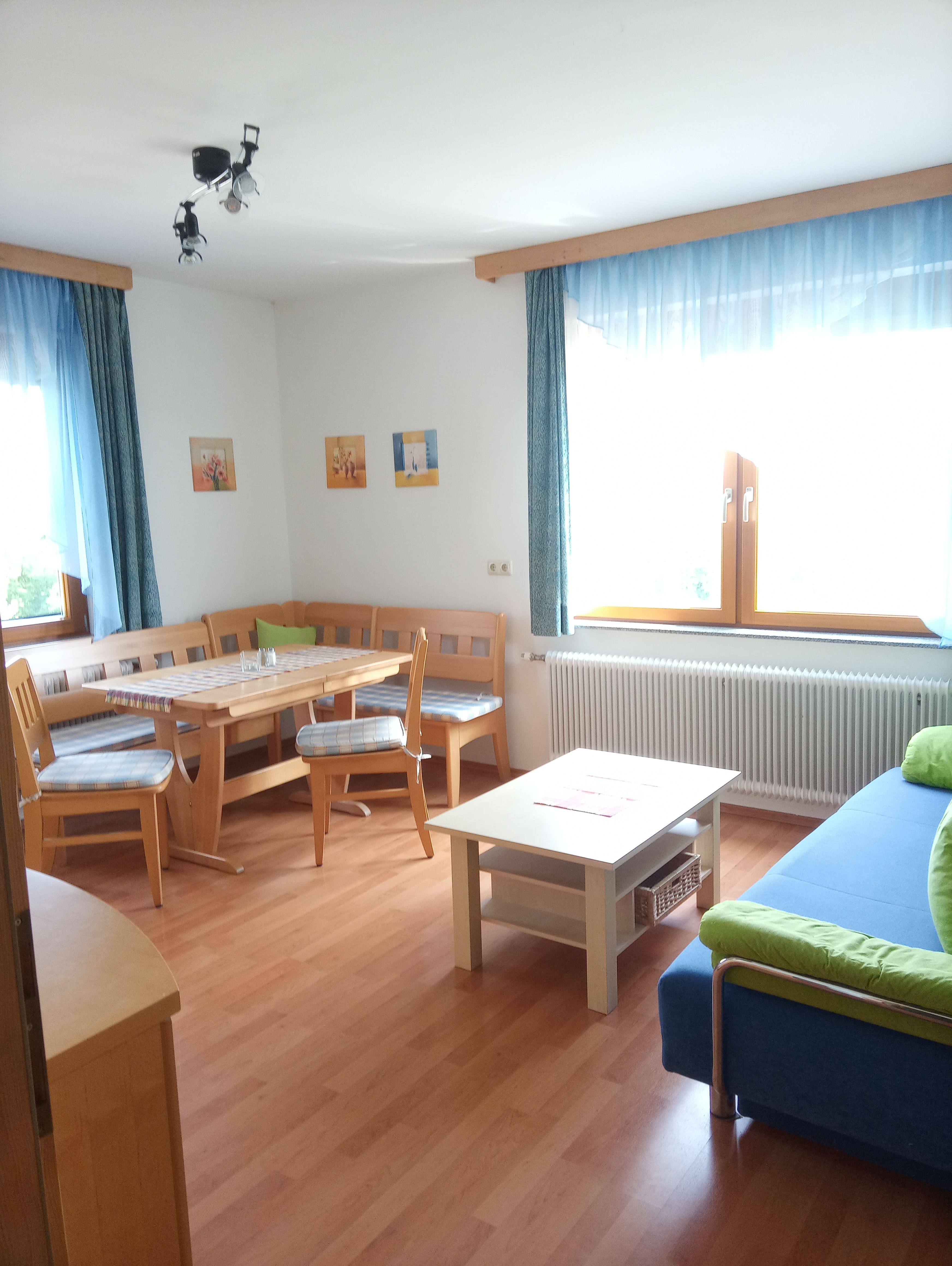 Světlý obývací pokoj s dřevěným nábytkem, rohovou lavicí, stolem, pohovkou a velkými okny s modrými závěsy.