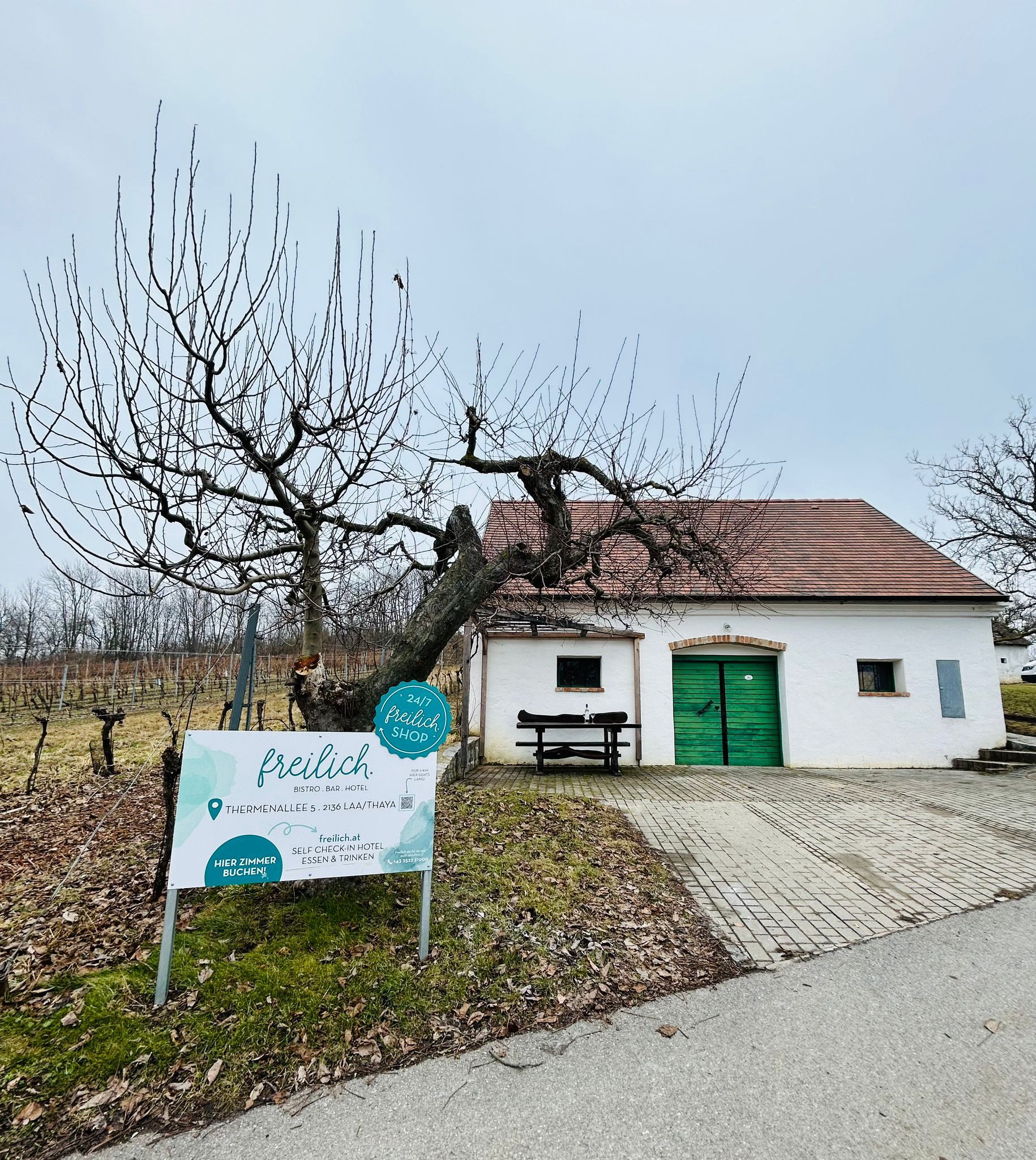 Bílá budova se zelenou bránou a nápisem "freilich" ve venkovském prostředí.