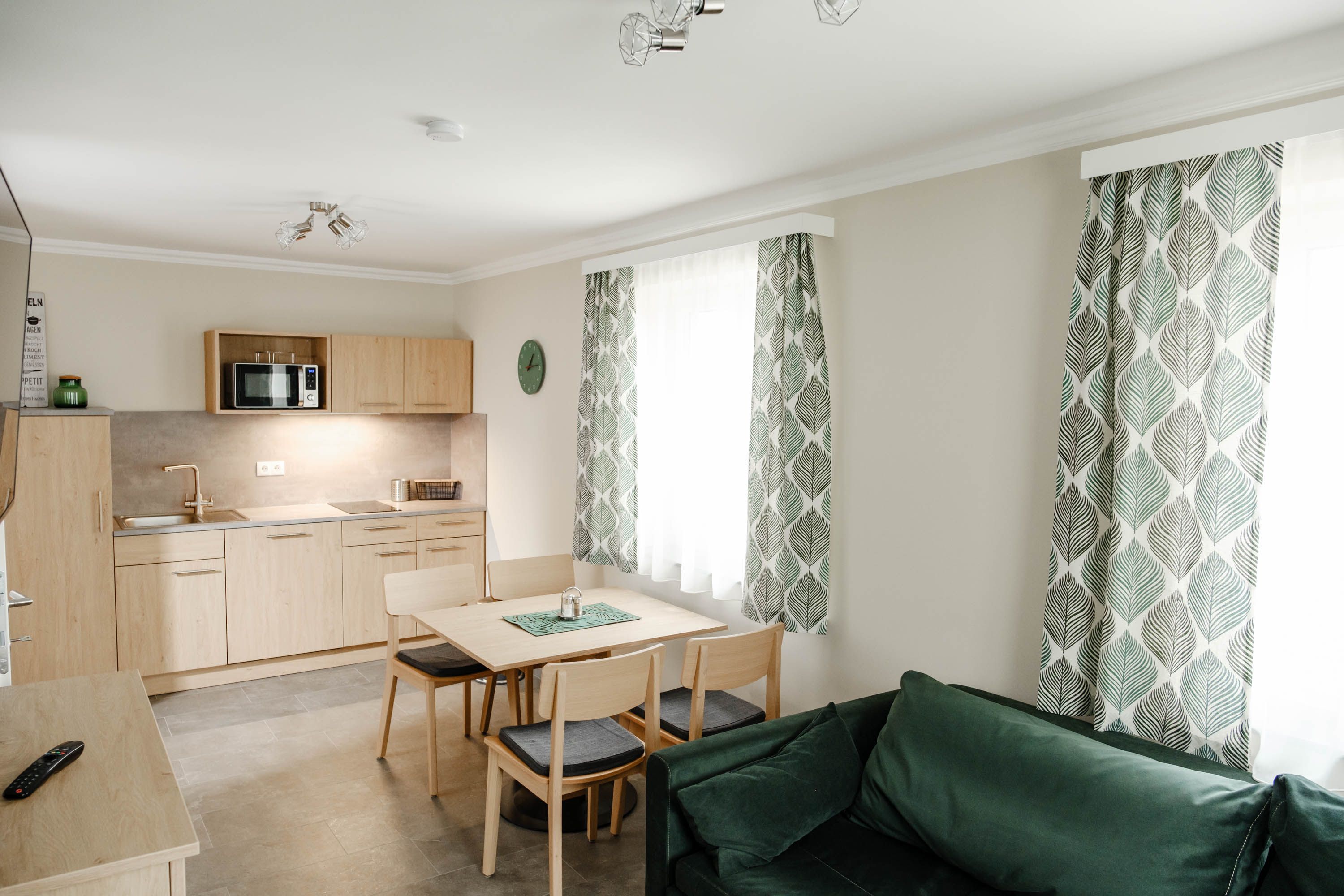Moderní kuchyně s dřevěným nábytkem, jídelním stolem, zelenými závěsy a pohovkou.