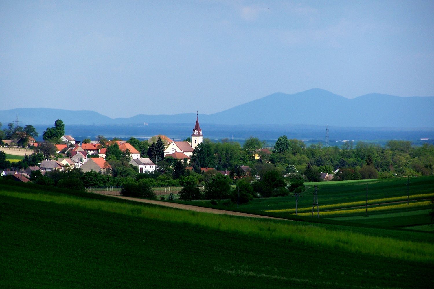 Krajina s vesnicí a kostelem, obklopená zelenými poli a kopci v pozadí.