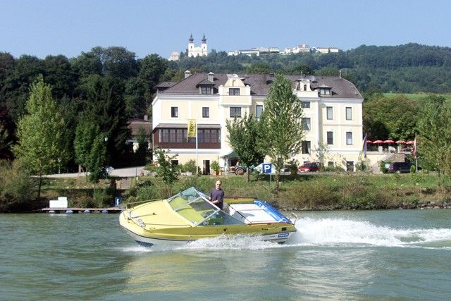 Žlutý motorový člun pluje po řece před velkou světlou budovou s kopci v pozadí.