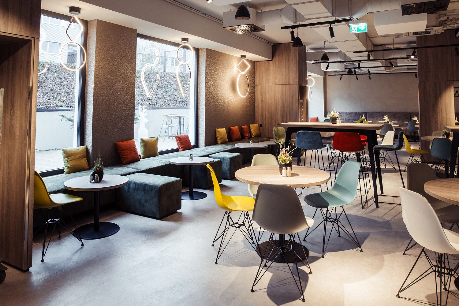 Moderní hotelová restaurace s barevnými židlemi a stoly, velkými okny a stylovým osvětlením.