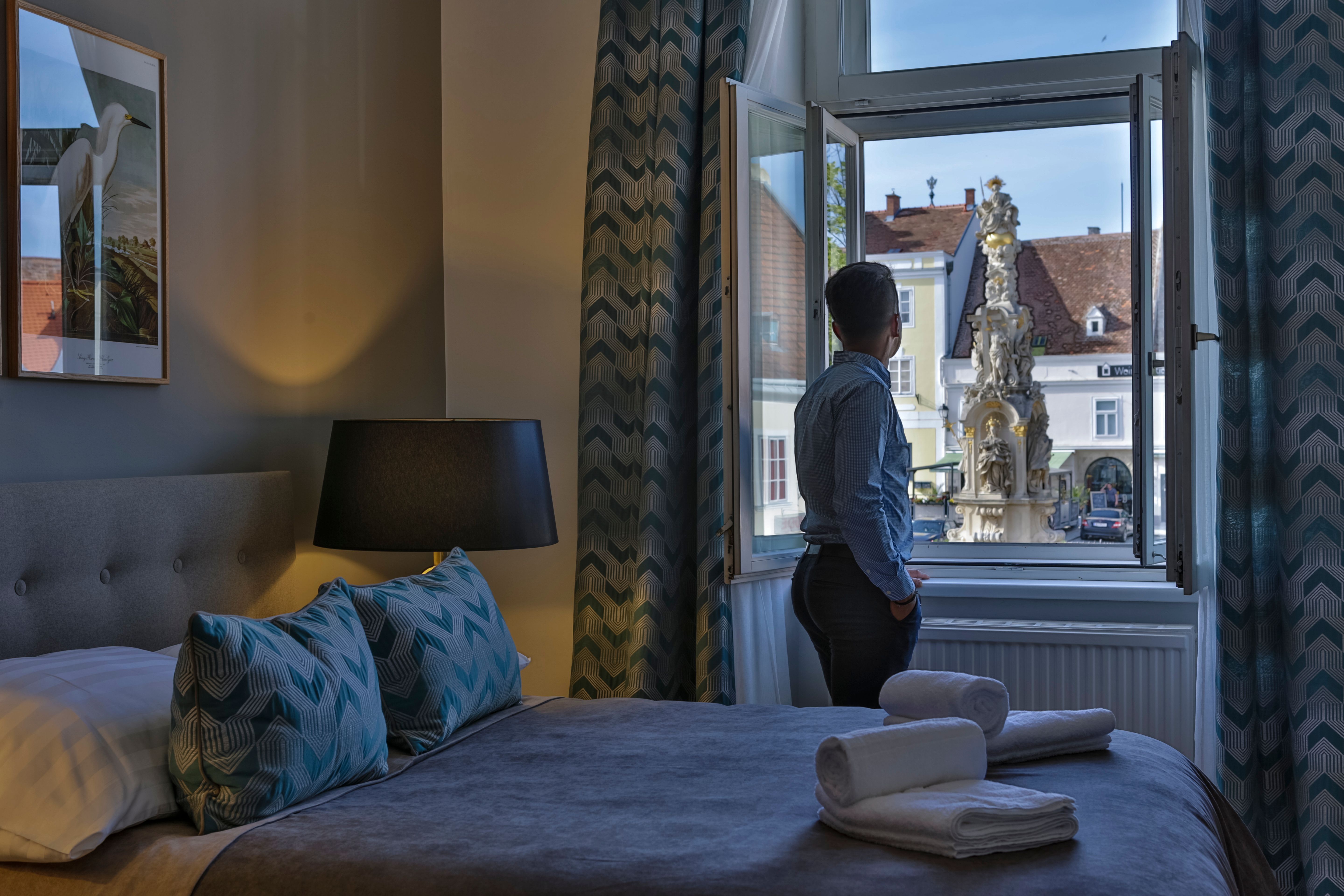 Osoba stojí u okna hotelového pokoje a dívá se na venkovní fontánu.