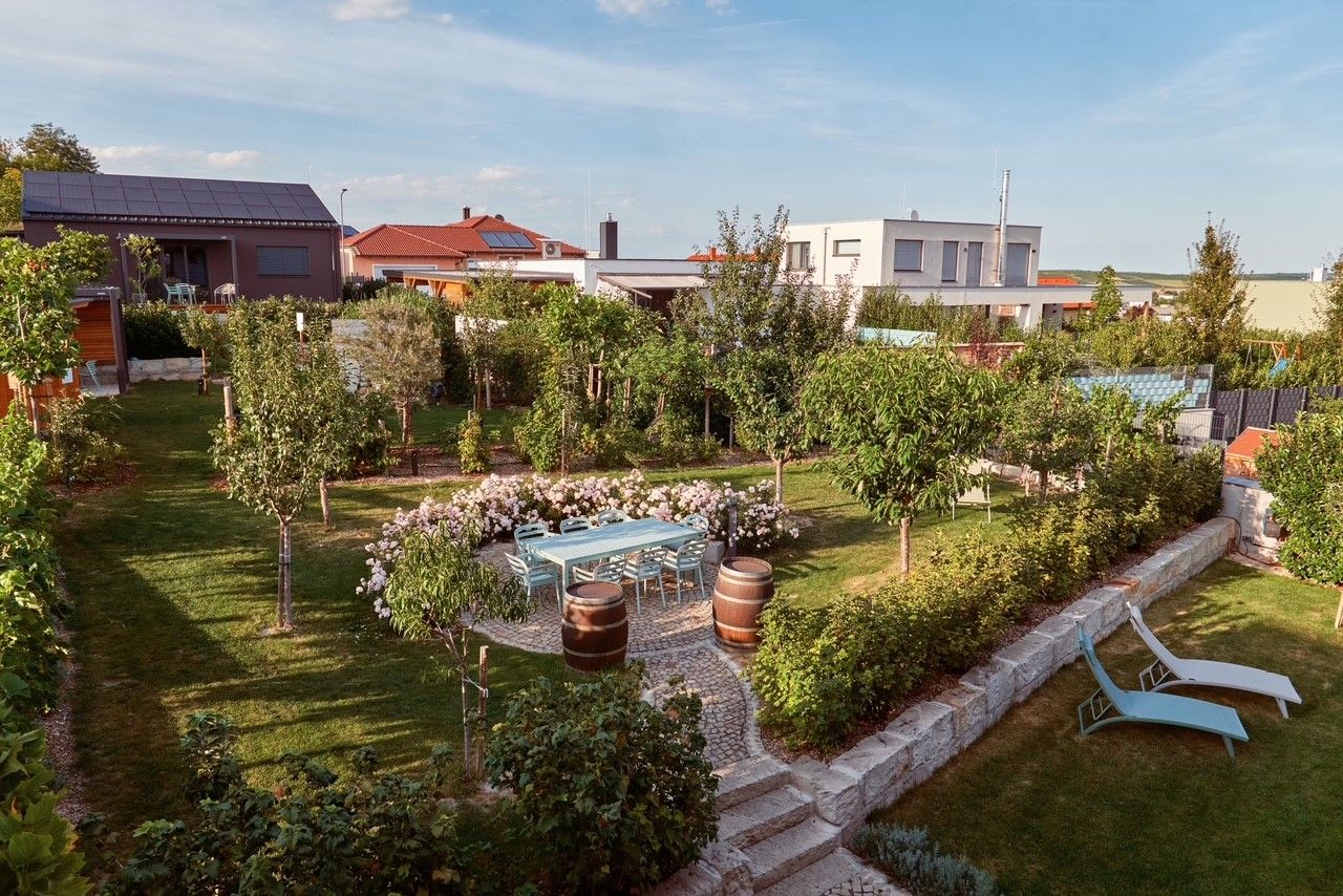 Udržovaná zahrada se stromy, květinami, stolem se židlemi a dvěma lehátky. V pozadí jsou vidět moderní domy.