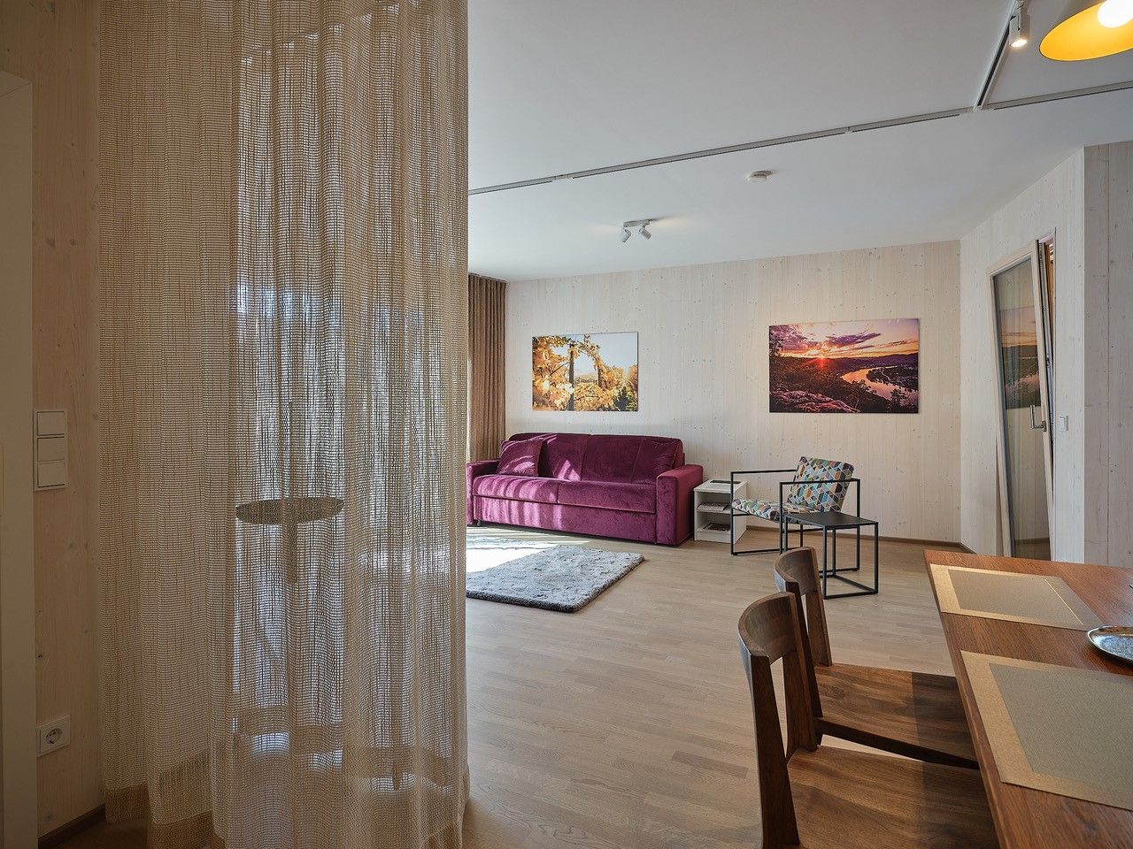 Obývací pokoj s fialovou pohovkou, dřevěným stolem a židlemi, závěsem a nástěnnými malbami.