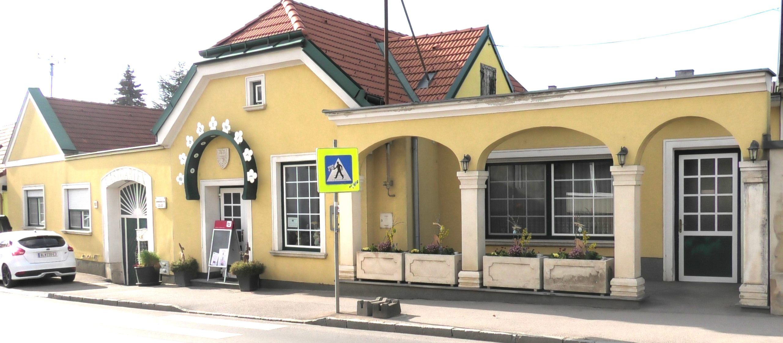 Pohled zvenčí na žlutou budovu s arkádami a červenou střechou.