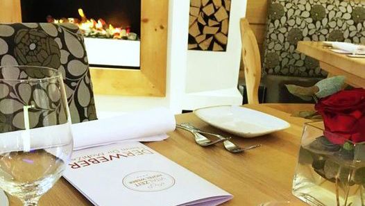 Pohled do interiéru restaurace s prostřeným stolem, sklenicí na víno, jídelním lístkem a krbem.