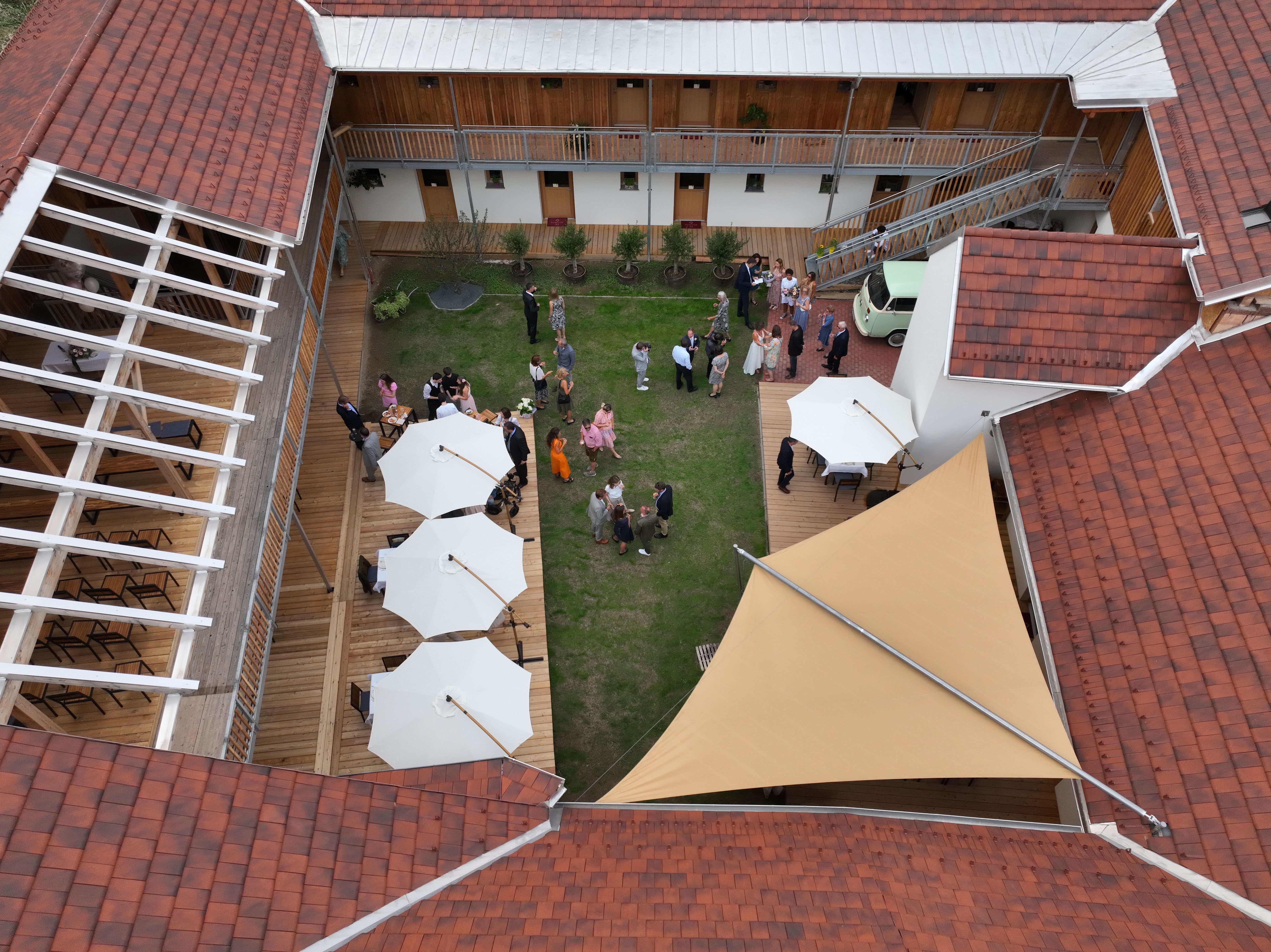 Pohled z ptačí perspektivy na vnitřní nádvoří se zeleným trávníkem, bílými slunečníky a hosty na recepci.