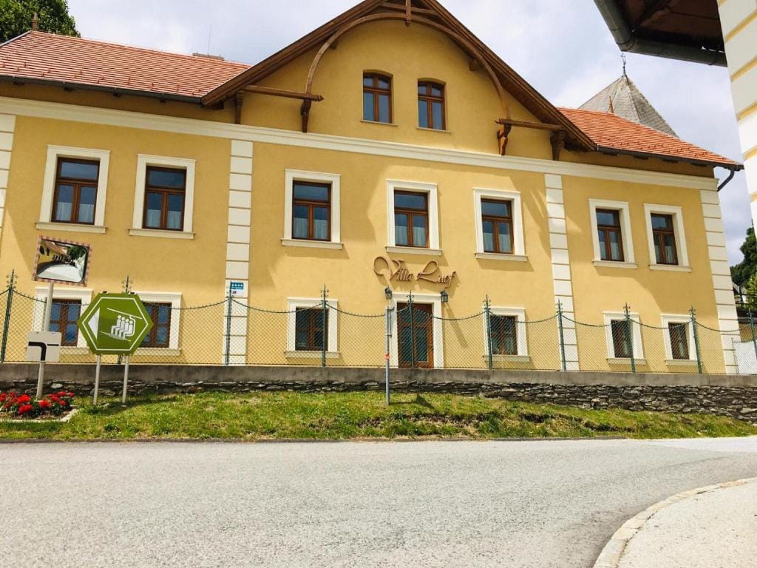 Žlutá budova s nápisem "Villa Luef", obklopená plotem a silnicí v popředí.