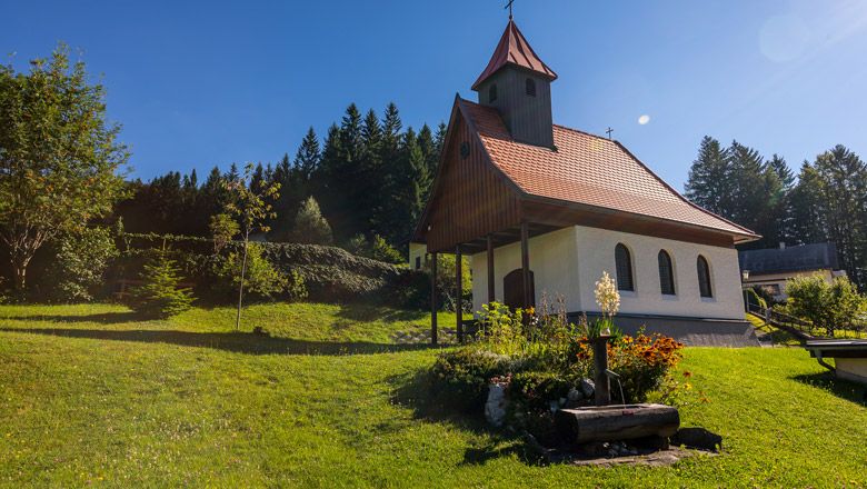 Malá kaple s červenou střechou na zelené louce, obklopená stromy a květinami.