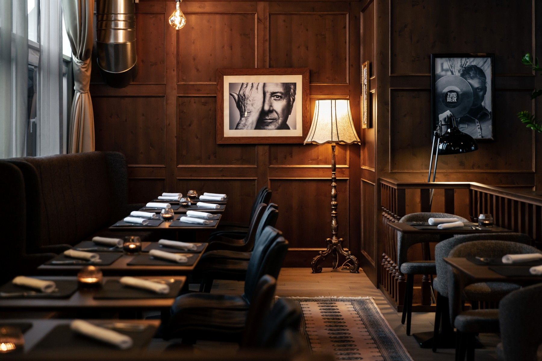 Útulný pokoj s dřevěnými stěnami, položenými stoly, stojací lampou a černobílými fotografiemi na stěnách.