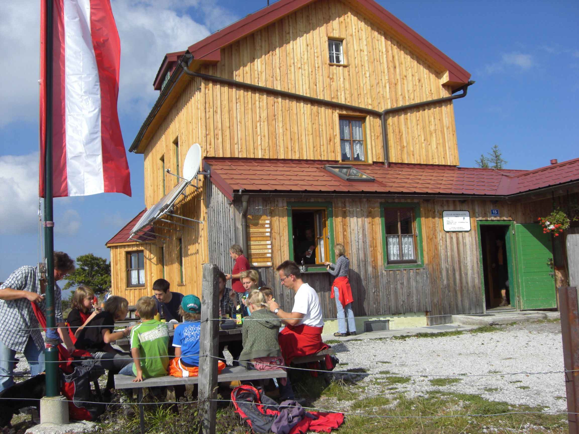 Skupina lidí sedí před horskou chatou s dřevěnou fasádou a rakouskou vlajkou.