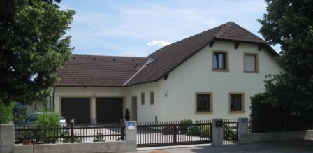 Moderní rodinný dům s hnědou střechou a světlým nátěrem, obklopený stromy a plotem.