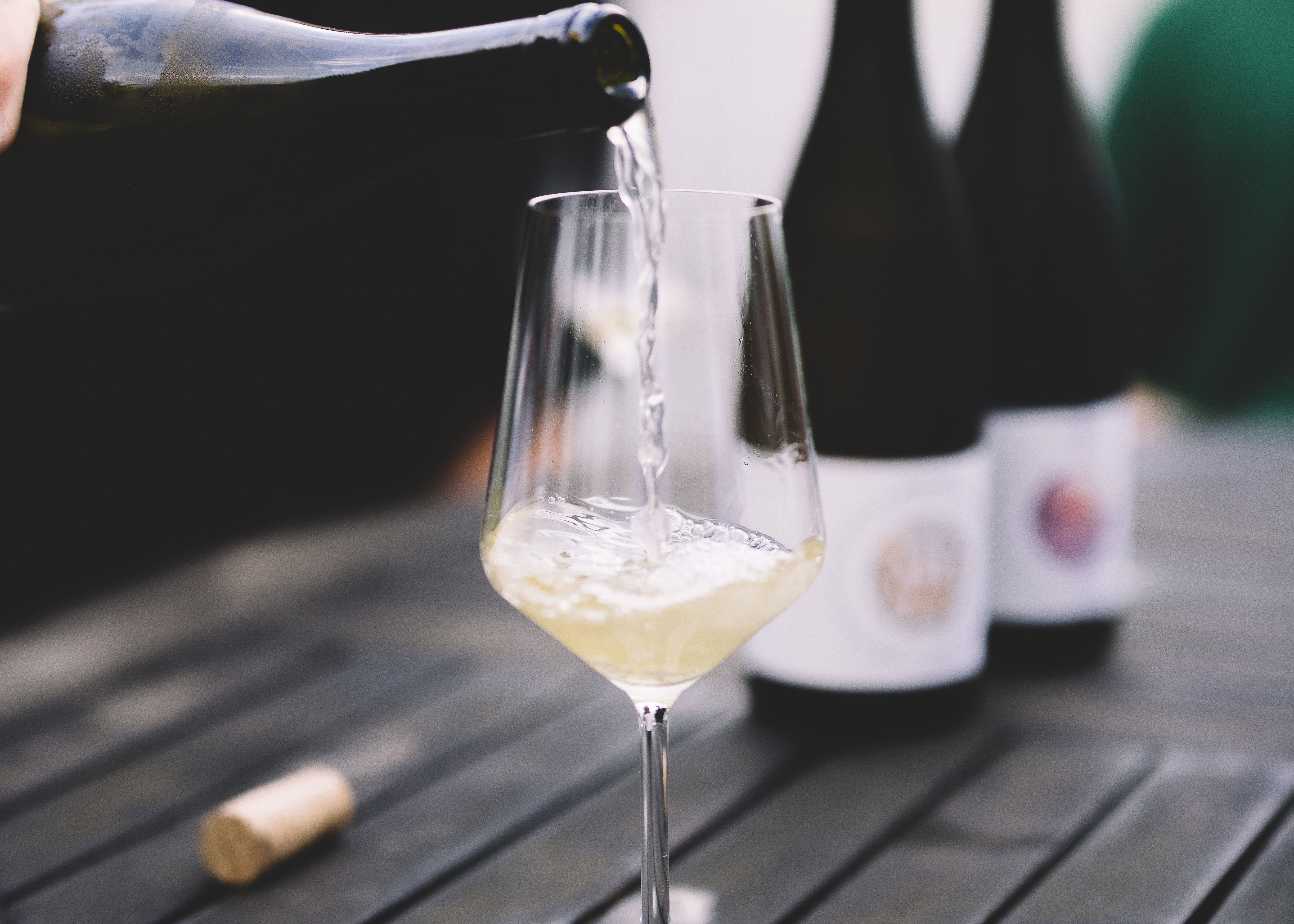 Na stole je nalita sklenice bílého vína a v pozadí jsou dvě láhve vína.