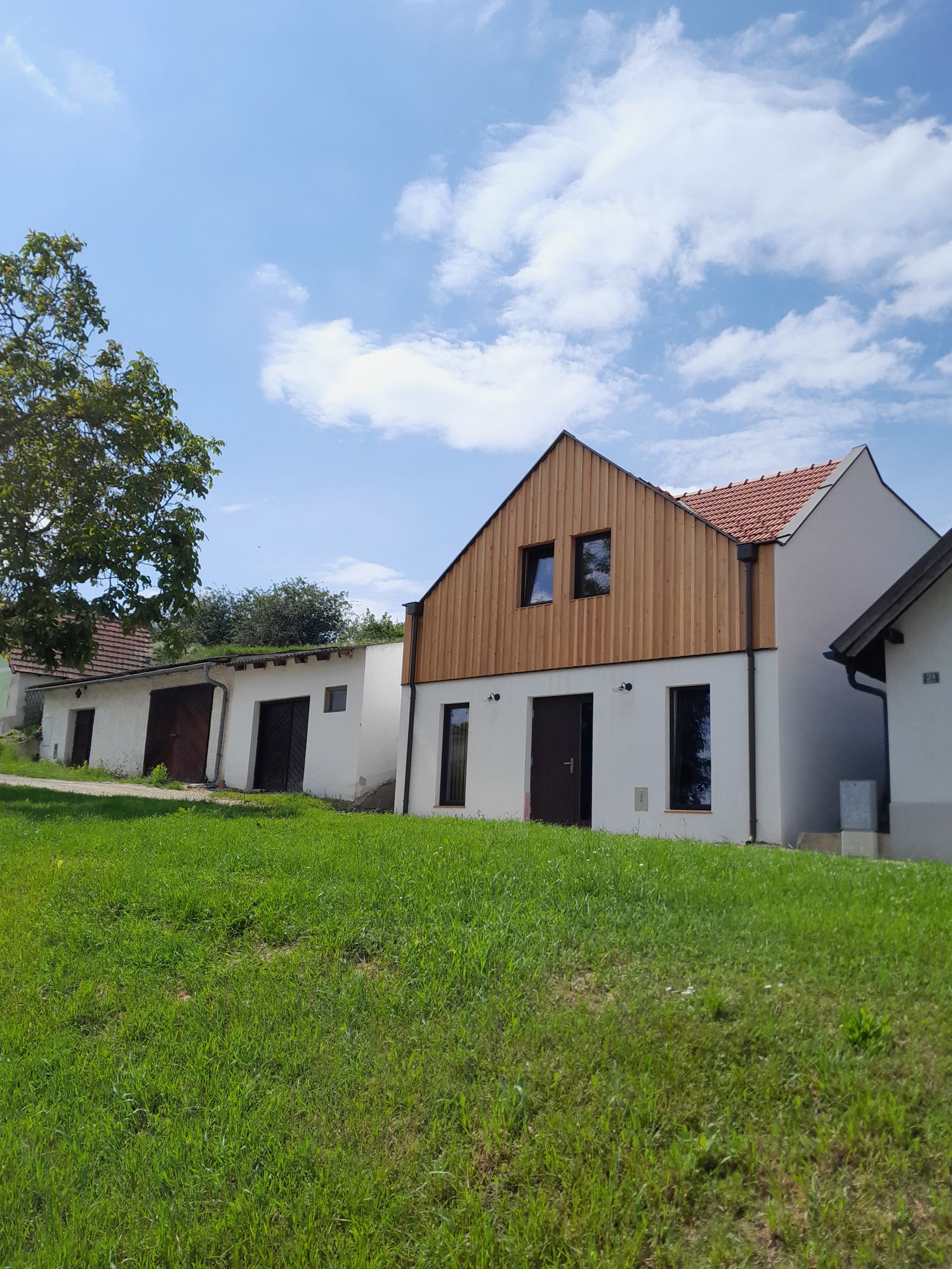 Na zeleném kopci pod modrou oblohou stojí moderní budova s dřevěnou fasádou a červenými střešními taškami.