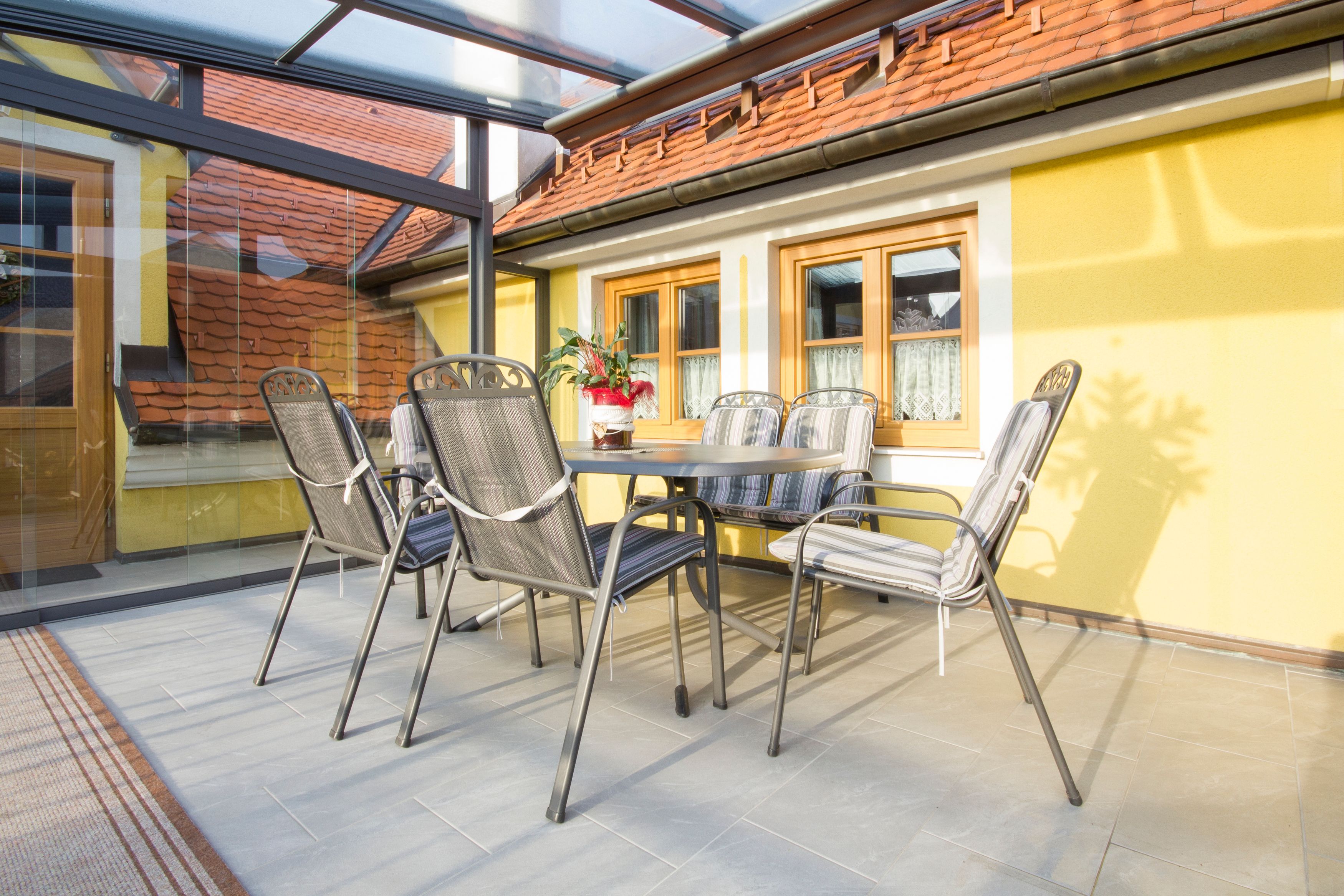 Zimní zahrada se stolem a židlemi, žluté stěny, skleněná střecha.