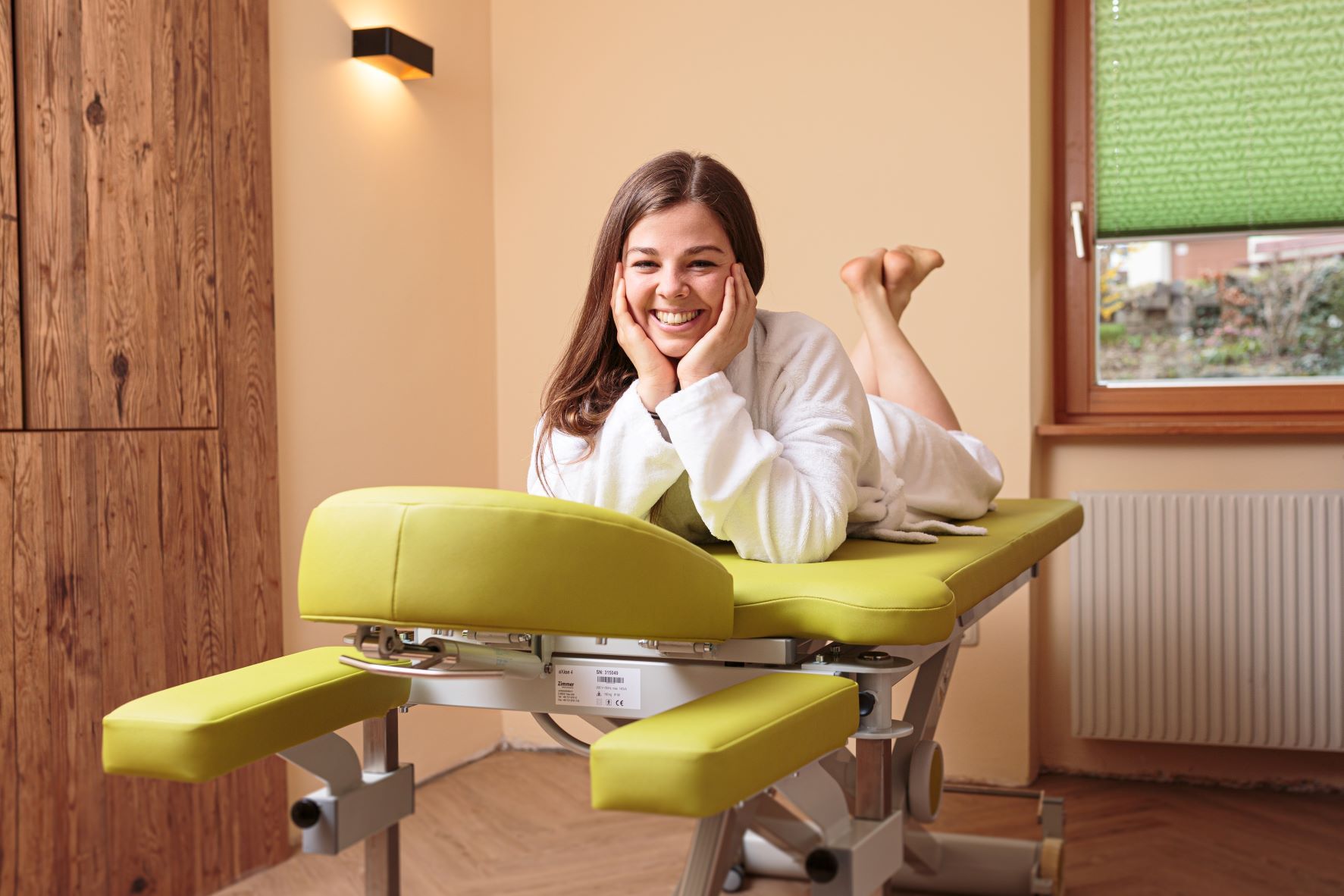 Žena v županu leží s úsměvem na zeleném masážním stole ve wellness místnosti.