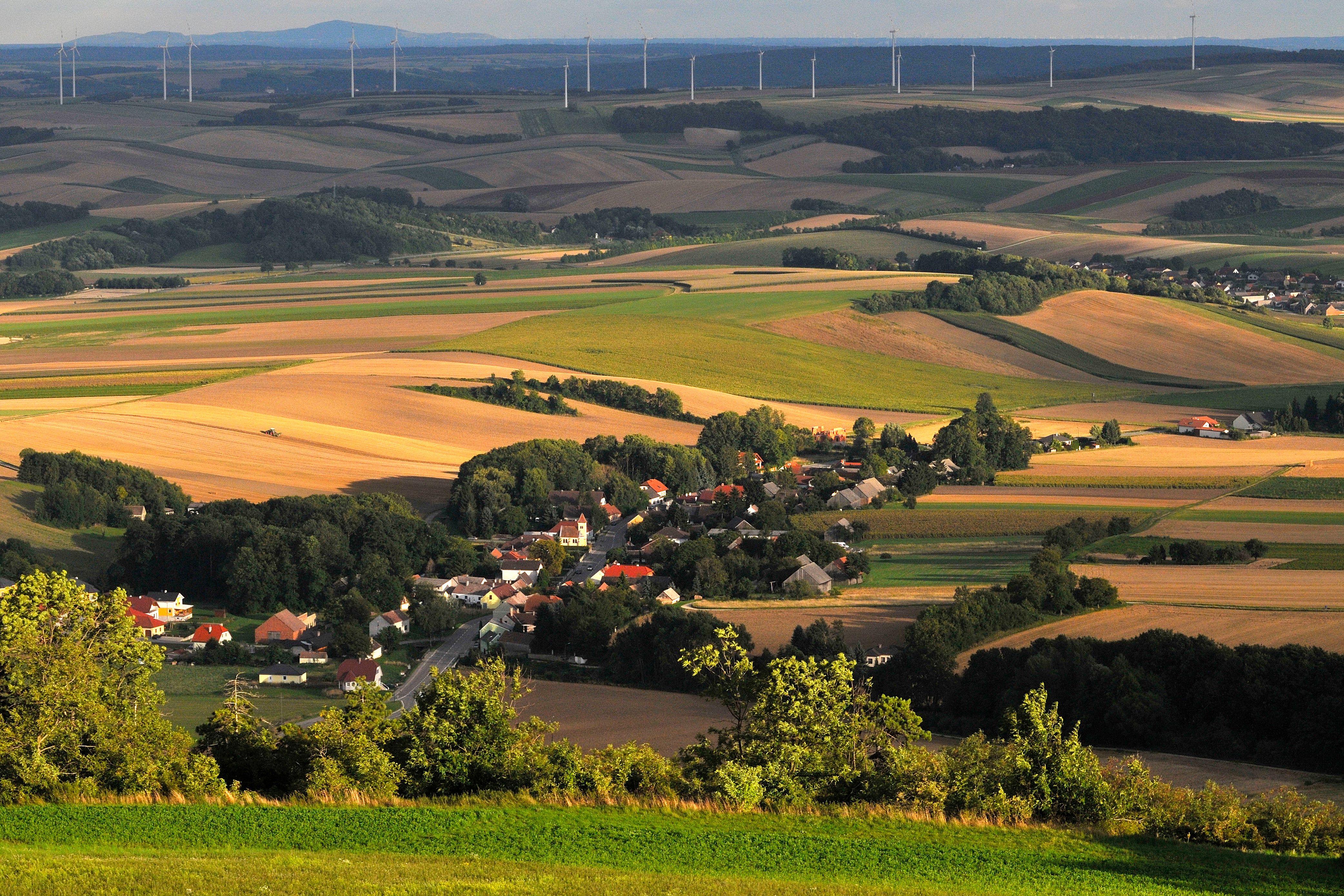 Krajina s poli, vesnicí a větrnými turbínami v pozadí.