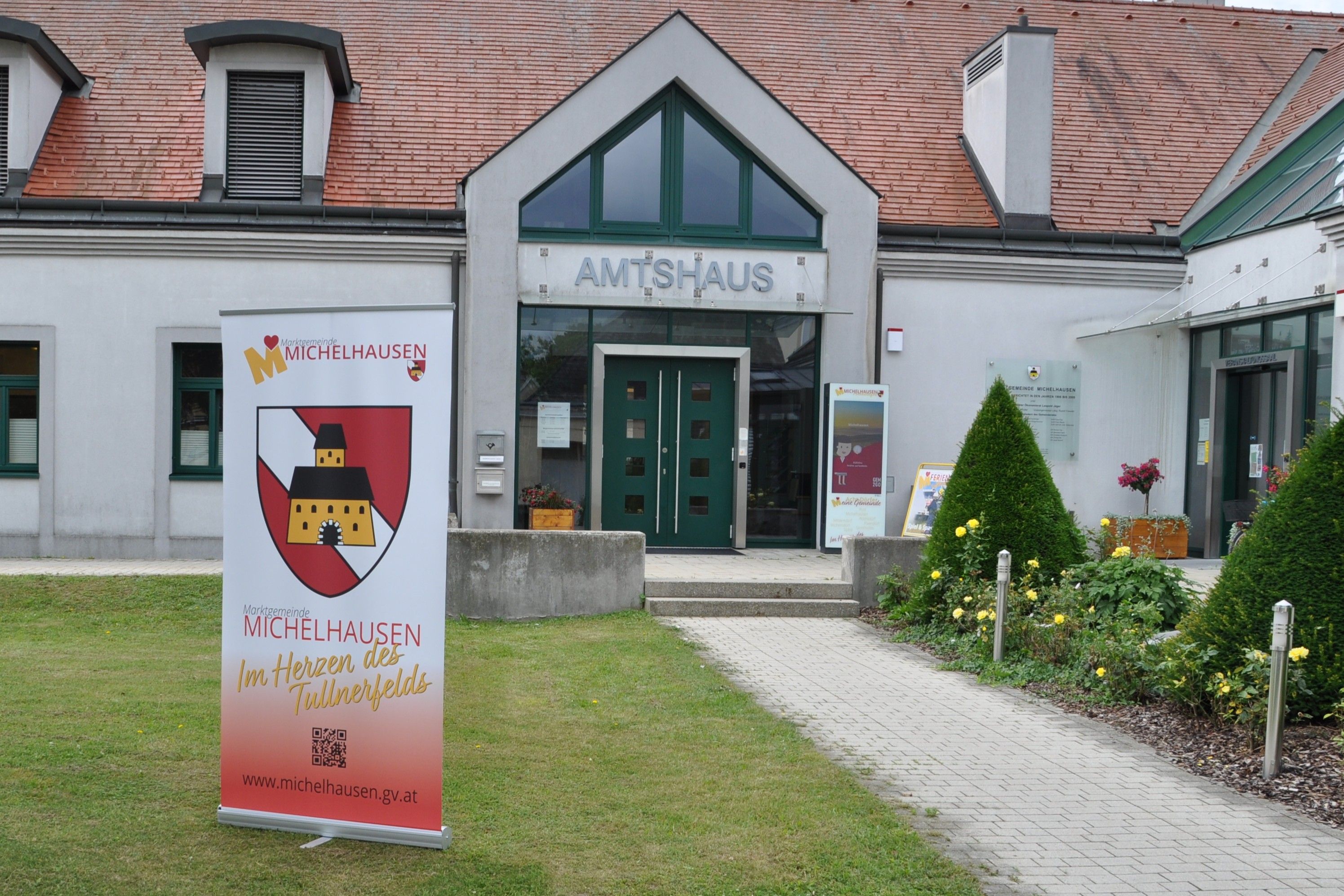 Vchod do Amtshausu Michelhausen s reklamním poutačem a upravenou zahradou.