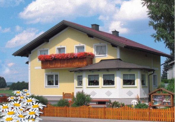Žlutý dům s červenou střechou a truhlíkem na květiny na balkoně, obklopený dřevěným plotem a kopretinami v popředí.