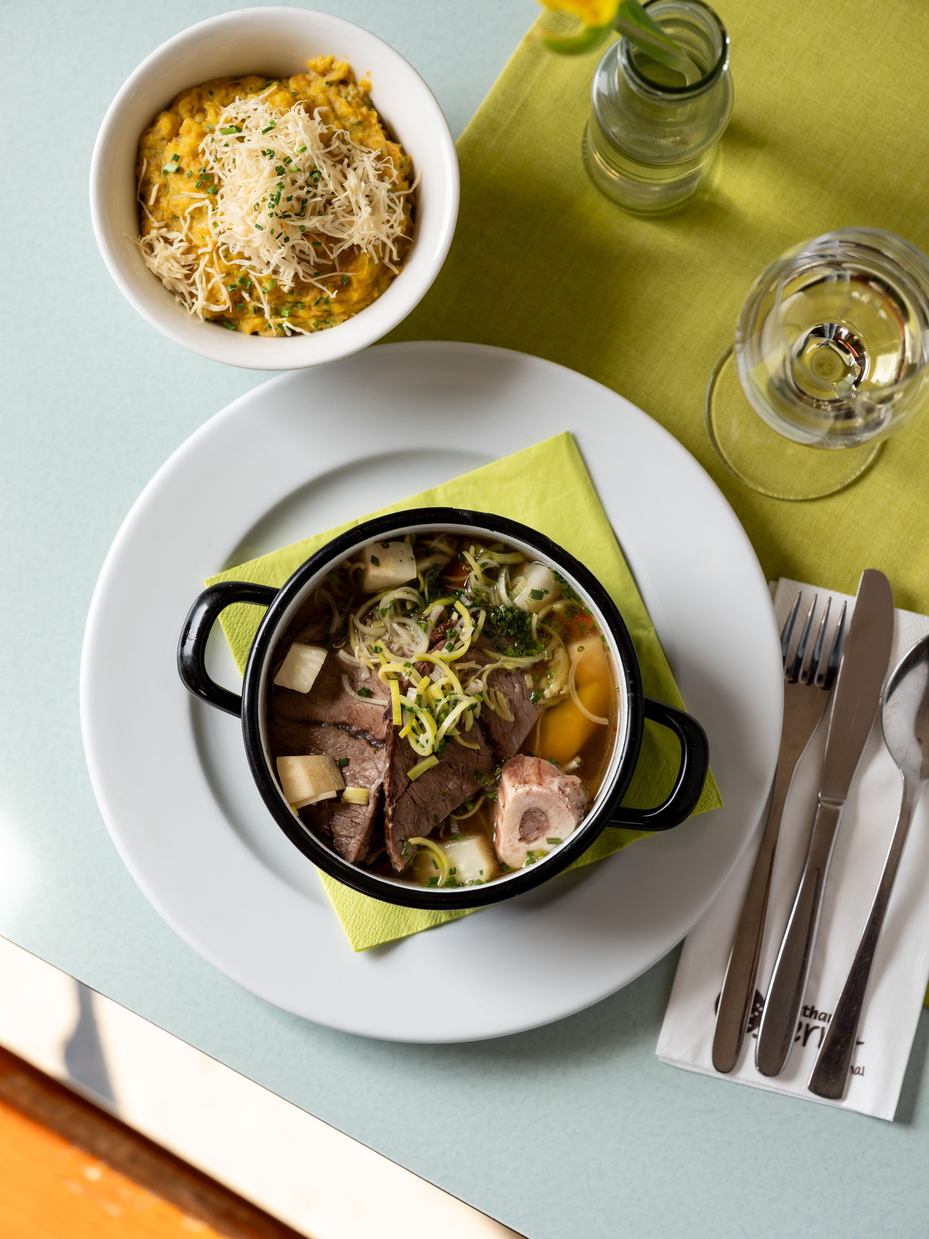 Stůl s talířem polévky, miskou ovesné kaše se sýrem, sklenicí vody a příborem na zeleném ubrousku.