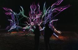 Zwei silhouettenhafte Personen stehen nachts vor einer großformatigen, abstrakten Lichtinstallation mit leuchtenden, organischen Formen in Violett-, Grün- und Blautönen; die Szene ist Teil des Lichtkunstfestivals „Blockheide leuchtet“.