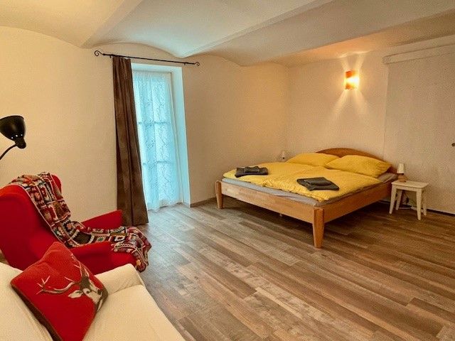 Útulná ložnice s dřevěnou podlahou, manželskou postelí, červeným křeslem a oknem se závěsem.