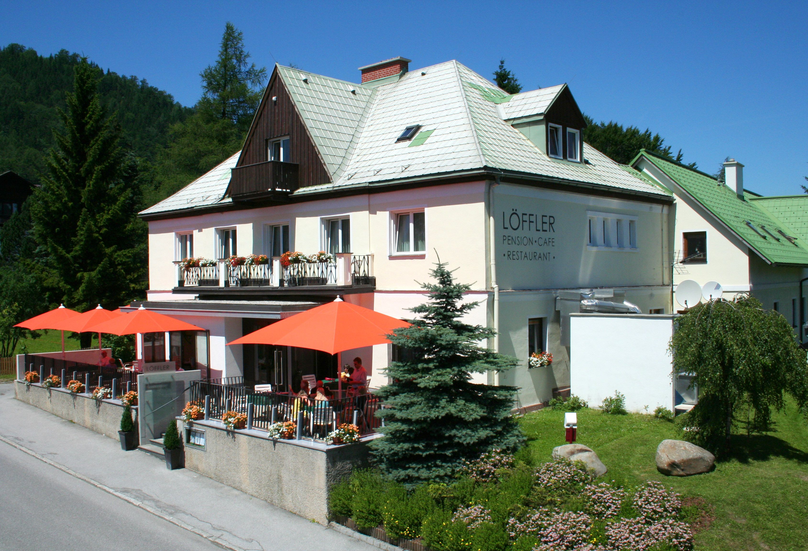 Dvoupatrová budova s nápisem "Löffler Pension-Café-Restaurant", obklopená stromy a květinami, s oranžovými slunečníky na terase.