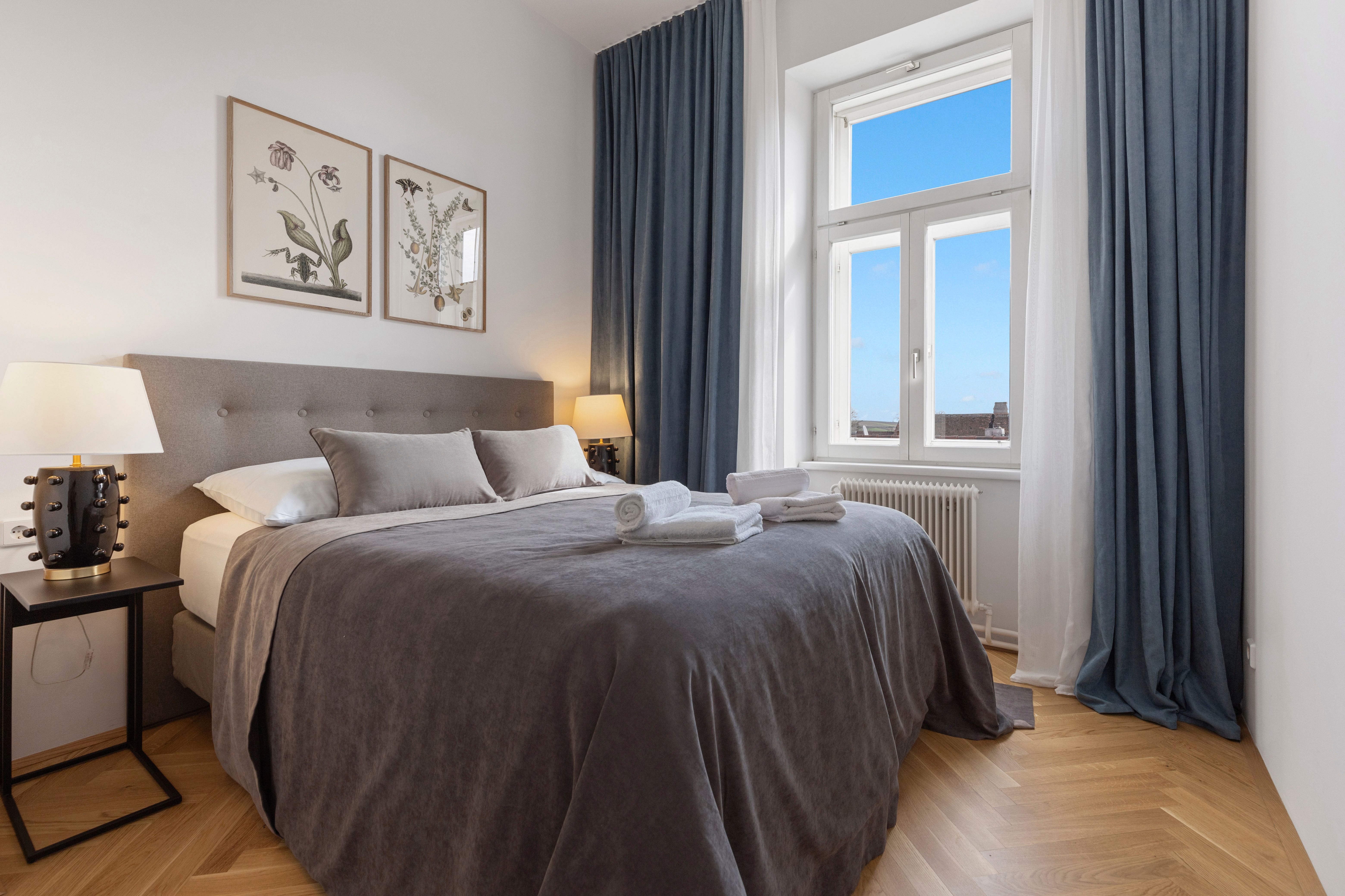 Elegantní ložnice s velkou postelí, šedým ložním prádlem, dvěma nočními stolky s lampičkami a oknem s modrým závěsem.