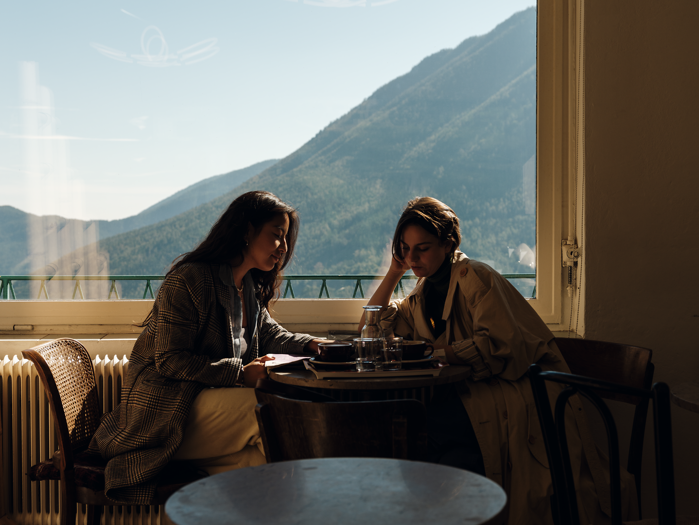 In der gemütlichen Atmosphäre der Kaffeehalle Semmering genießen zwei Freundinnen einen entspannten Moment mit Blick auf die majestätischen Berge. Das sanfte Licht, das durch die großen Fenster strömt, schafft eine einladende Stimmung, während sie sich über ihre Erlebnisse austauschen.