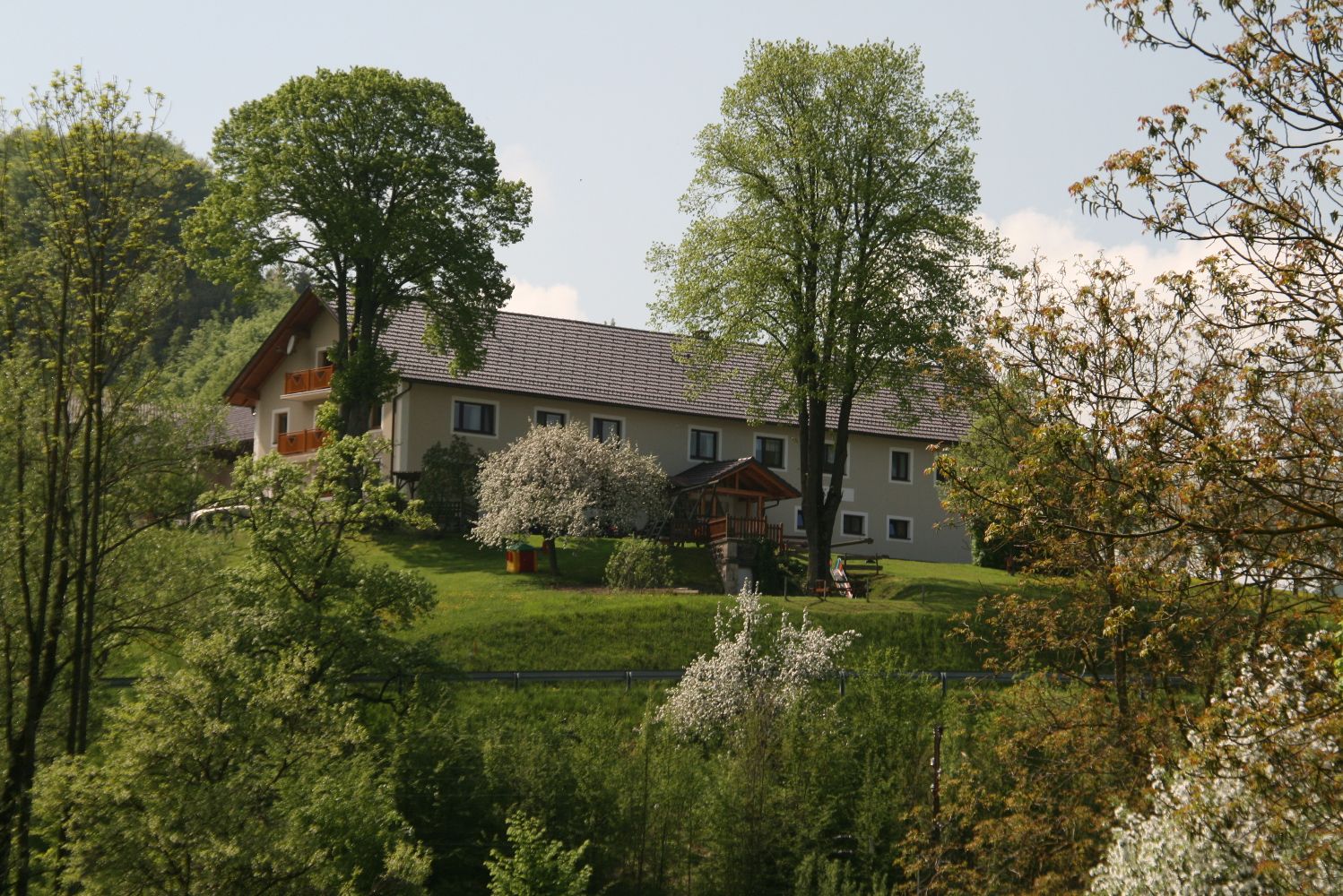 Farma na zeleném kopci, obklopená stromy a kvetoucími keři.