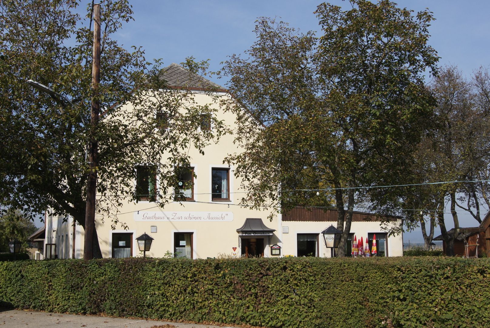 Žlutý hostinec s nápisem "Gasthaus Zur schönen Aussicht", obklopený stromy a živým plotem.