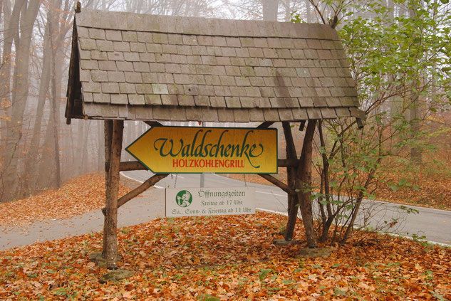 Dřevěná cedule v lese ukazuje na Waldschenke Holzkohlengrill.